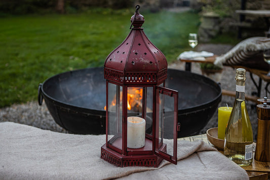 Red lantern with a candle on a table outdoors near a fire pit and wine bottle.