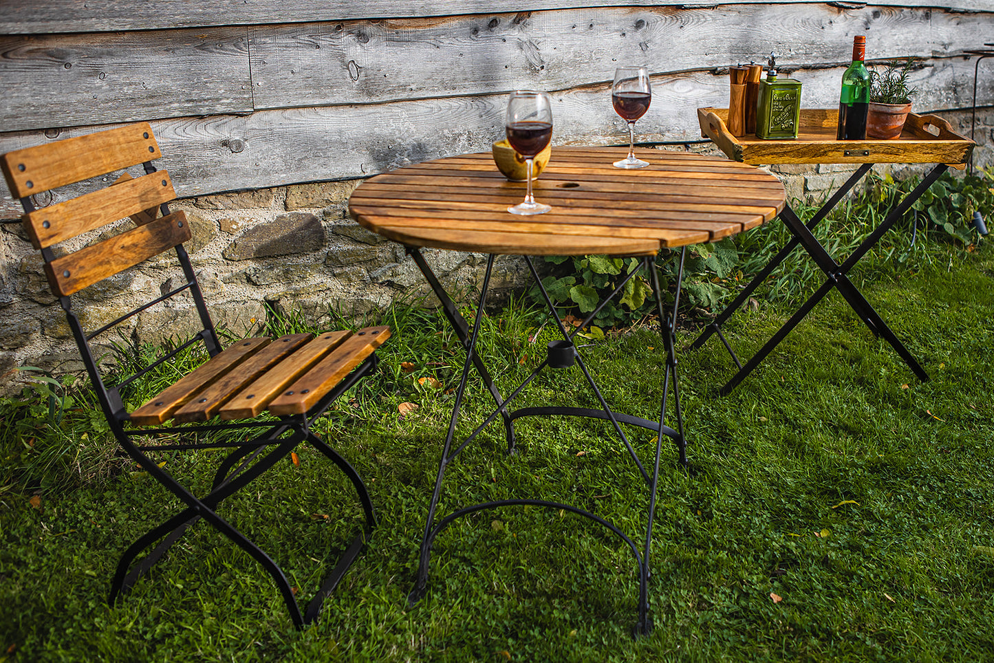 Garden table with chairs and wine glasses on a grassy area with a rustic wooden wall background.