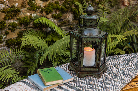 Decorative lantern with a candle on a table outdoors, surrounded by greenery.