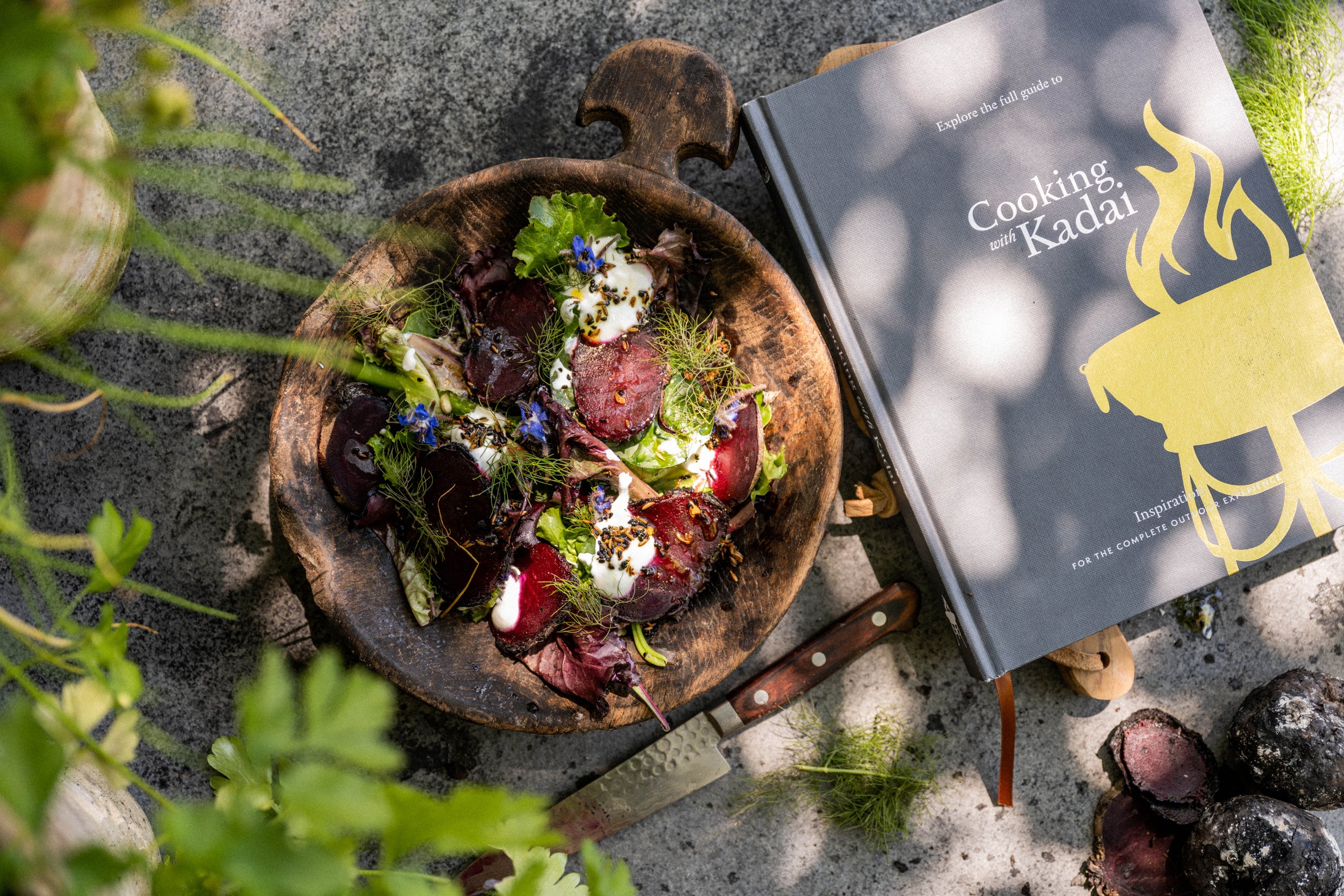 A wooden bowl with a salad of leafy greens, roasted beets, cream, and herbs, placed next to a cookbook titled “Cooking with Kadai.