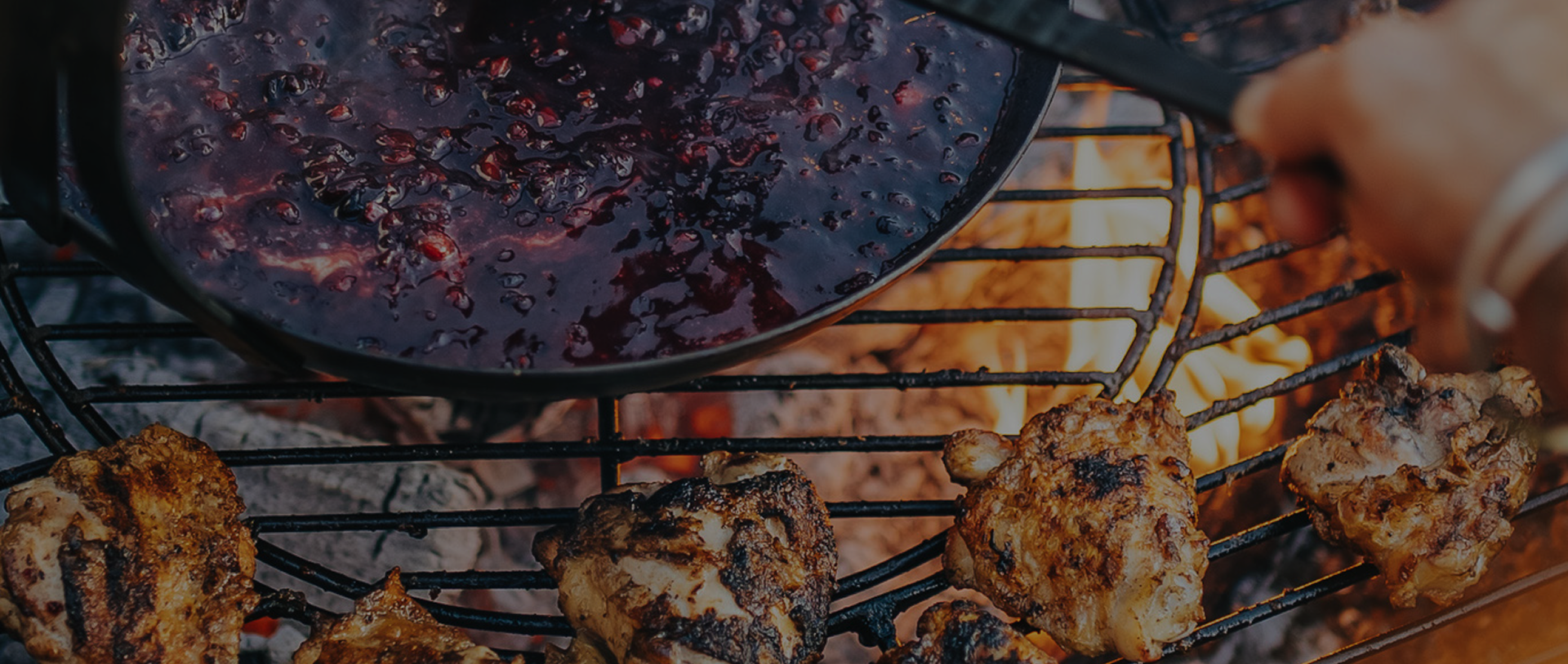 Grilled chicken cooking beside a pan of berry sauce over flames.