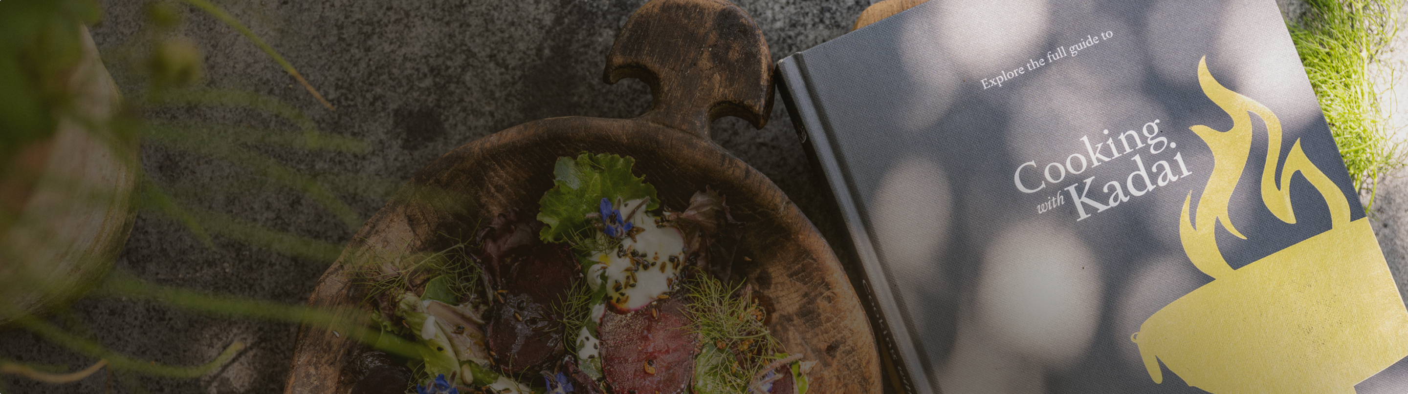 Wooden bowl with a colorful salad next to a cookbook titled "Cooking with Kadai" on a sunlit table.