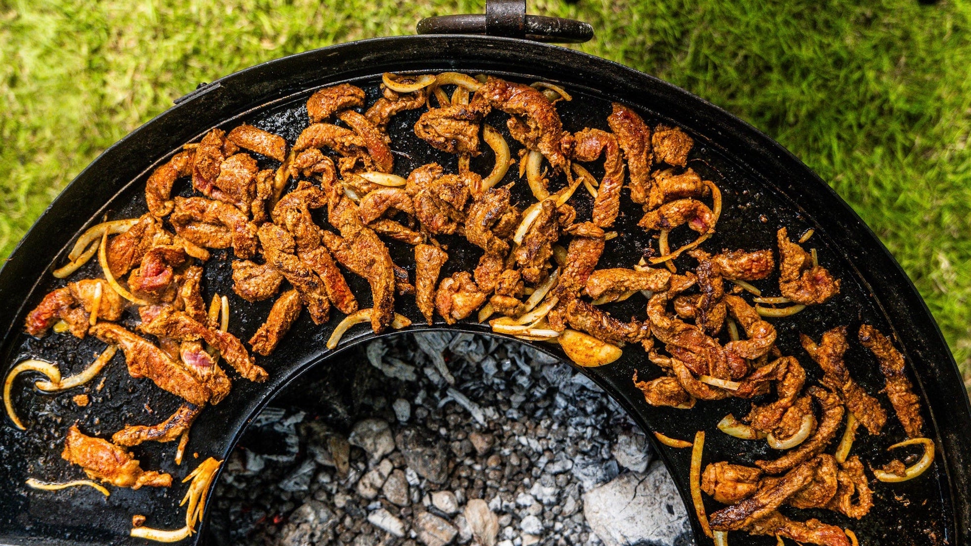 Thinly sliced BBQ beef shawarma cooking over embers and hot coals on a round BBQ grill.