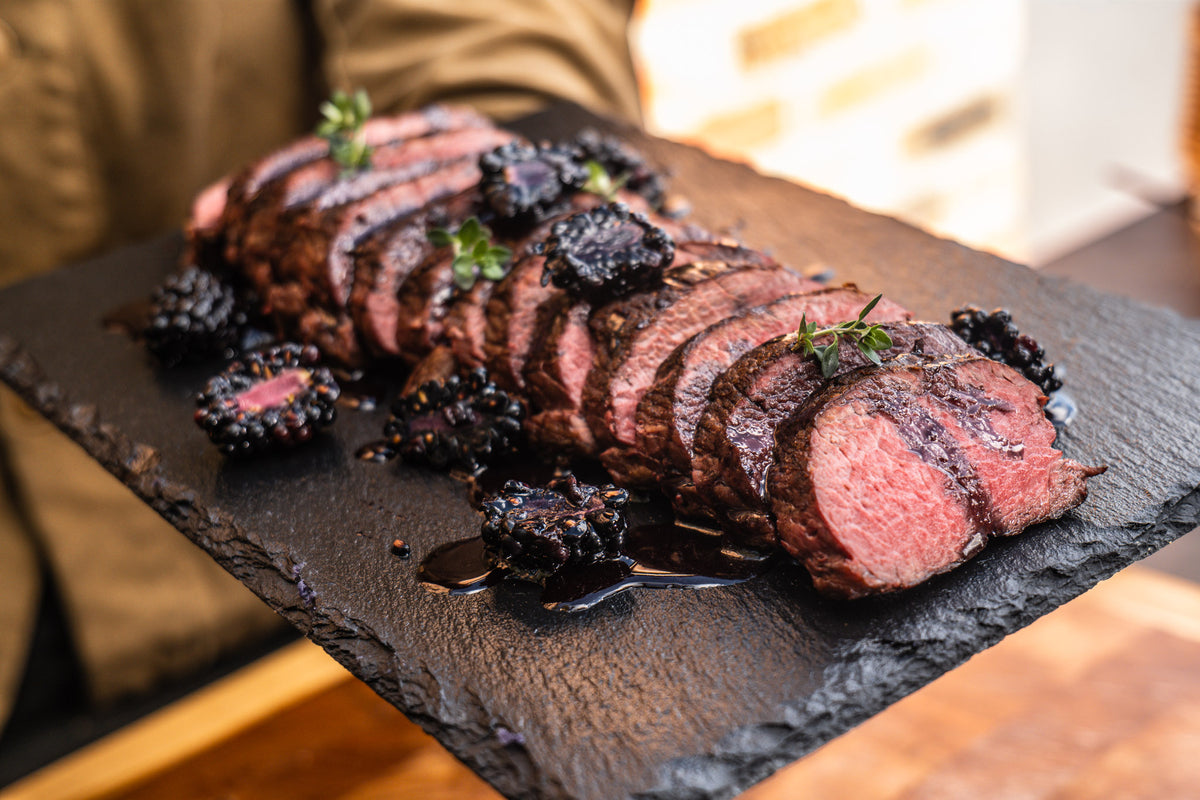 Sliced blackberry grilled venison tenderloin on a dark slate serving platter, garnished with fresh herbs and whole blackberries, held by a person in a kitchen setting.