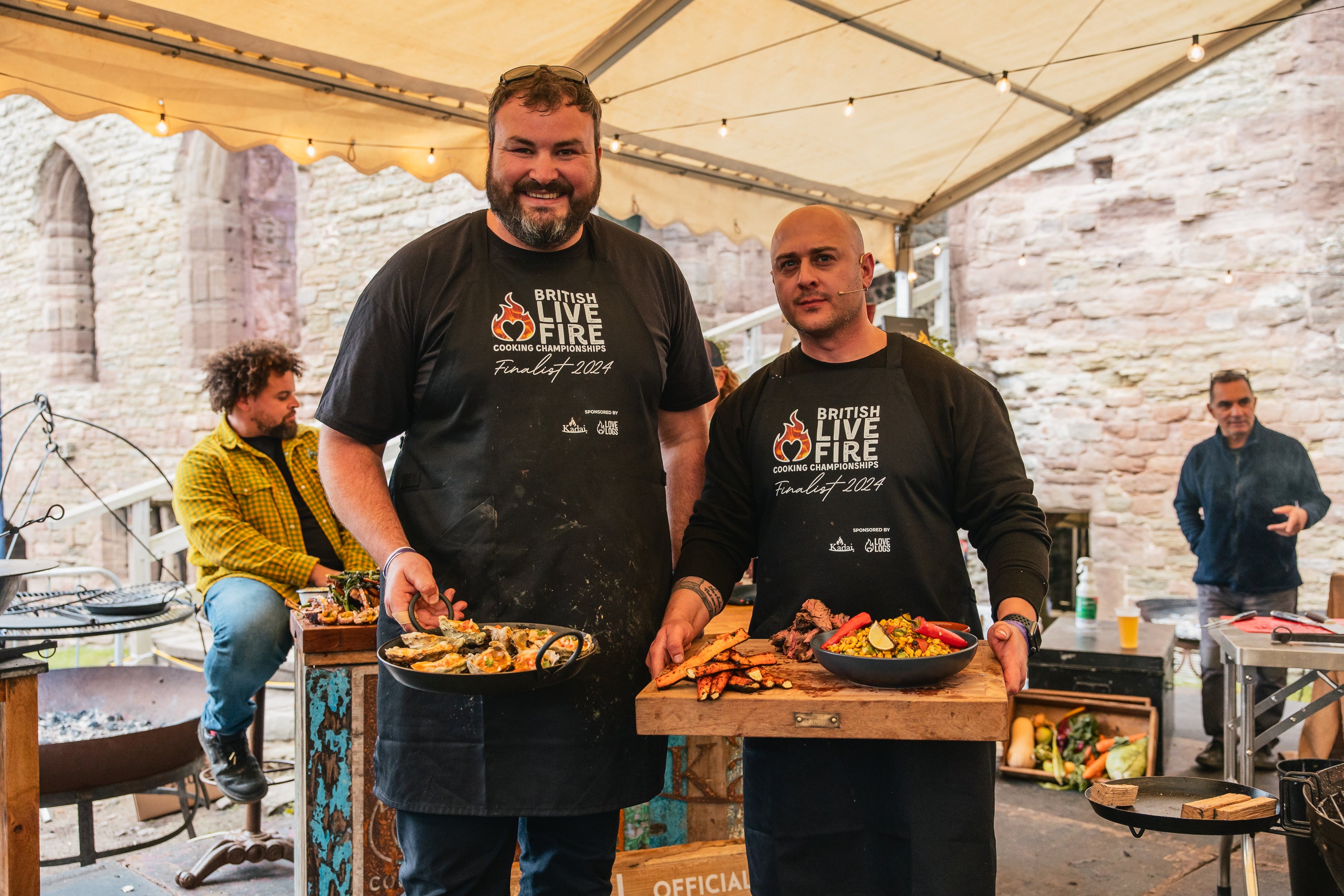 BLFCC Two men holding plates of food in a rustic outdoor setting with brick walls and wooden tables.