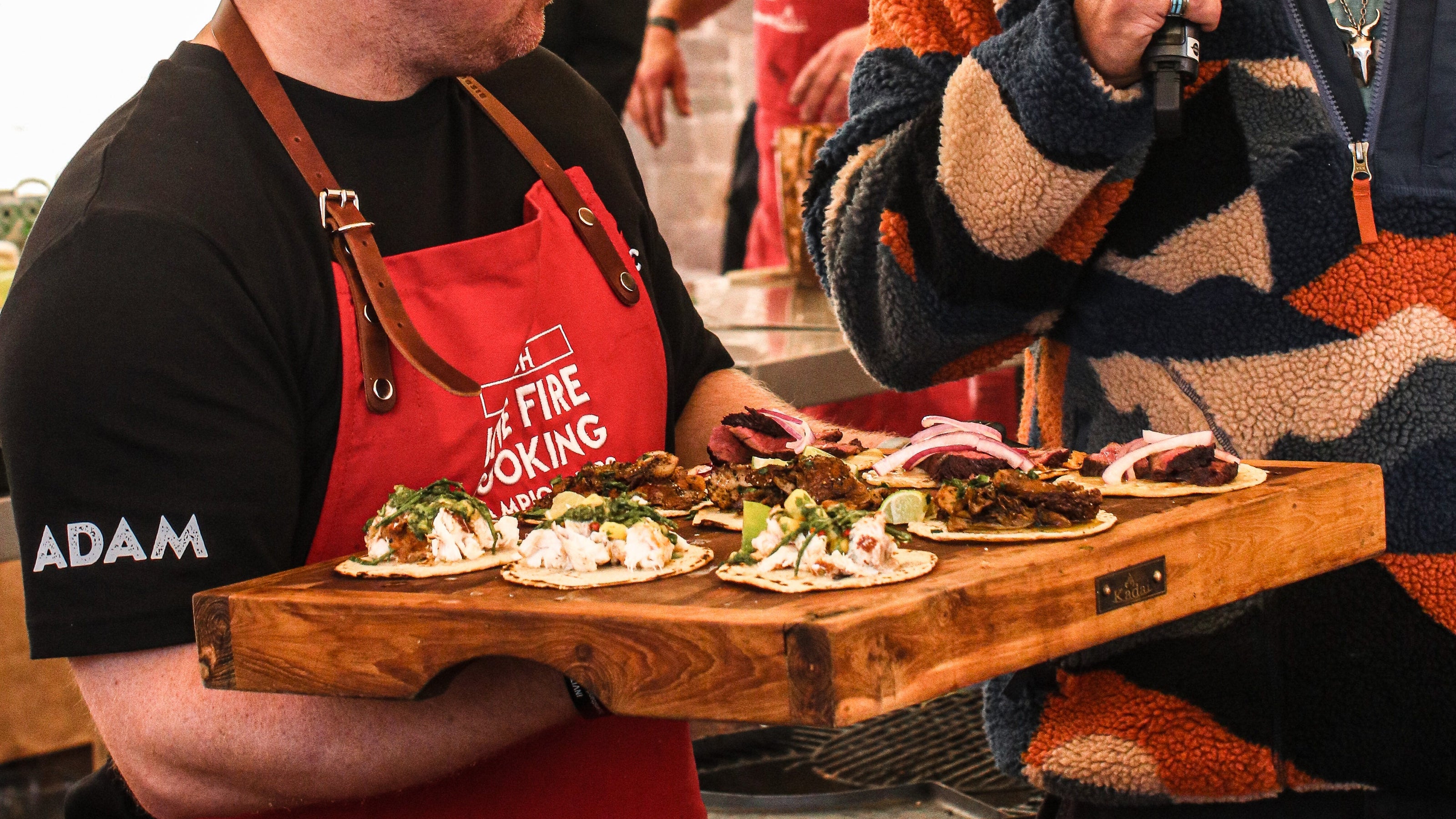 Man in a red apron holding a wooden board with food, speaking into a microphone.