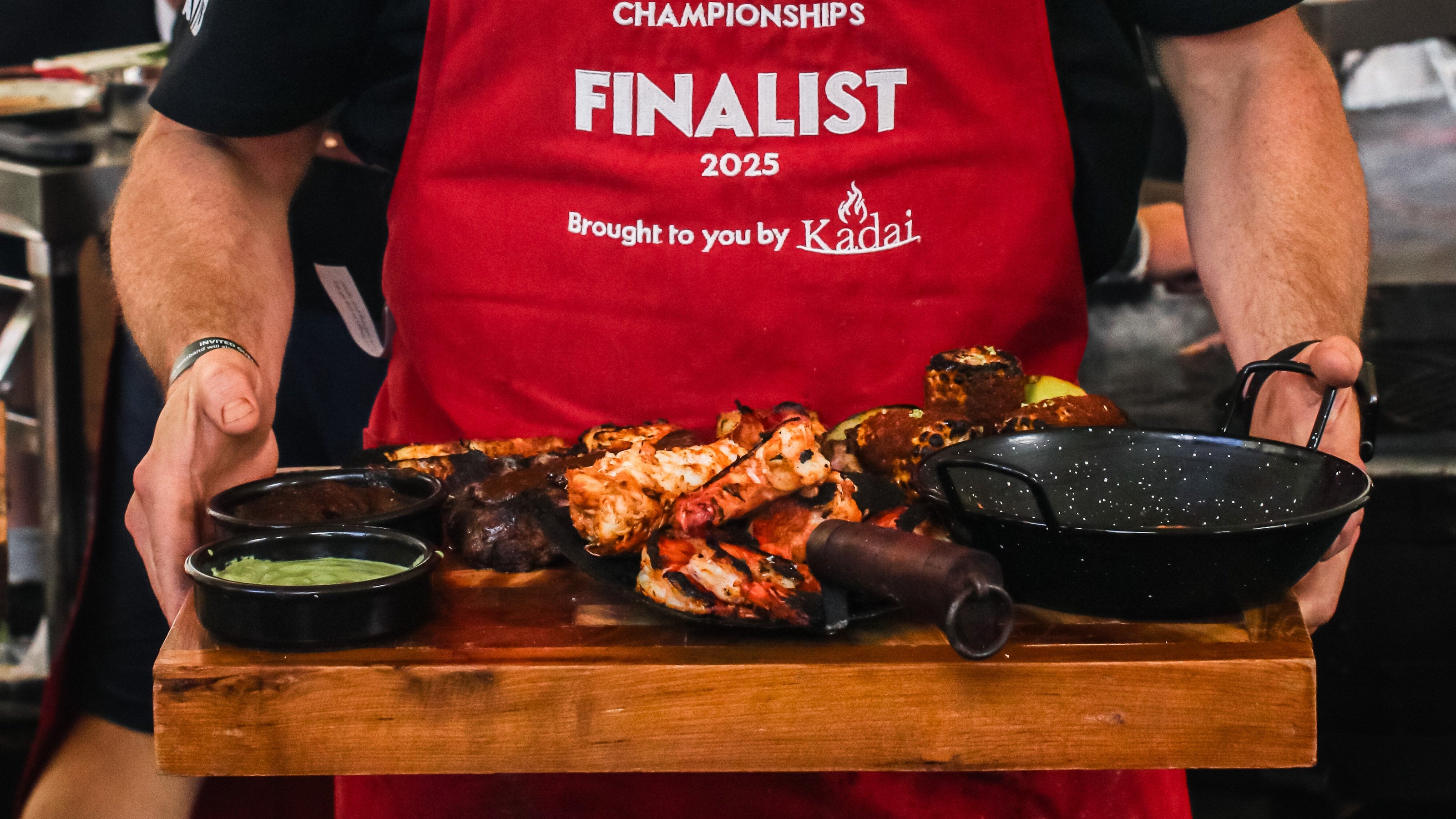 Man in a red apron holding a tray of food with 'Live Fire Cooking Championships Finalist' text.
