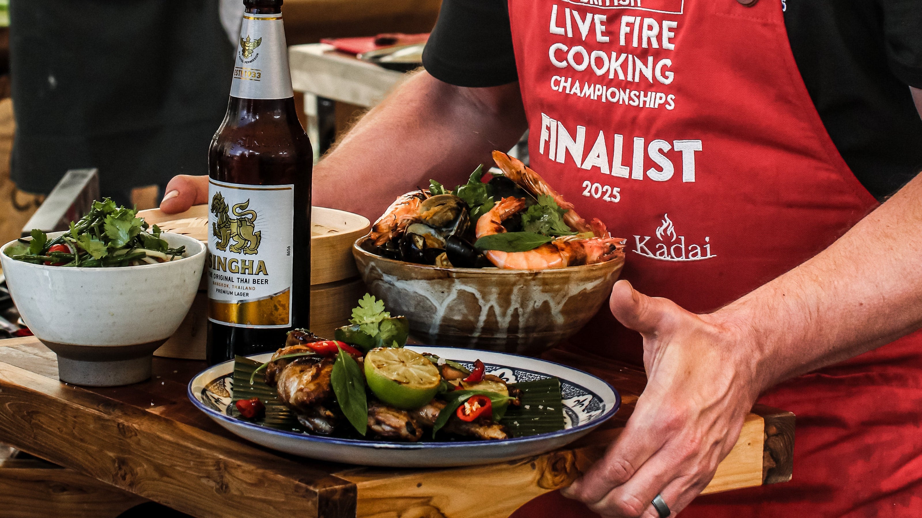 Man in a red apron holding a plate of food at a cooking event with 'Kadai' branding.