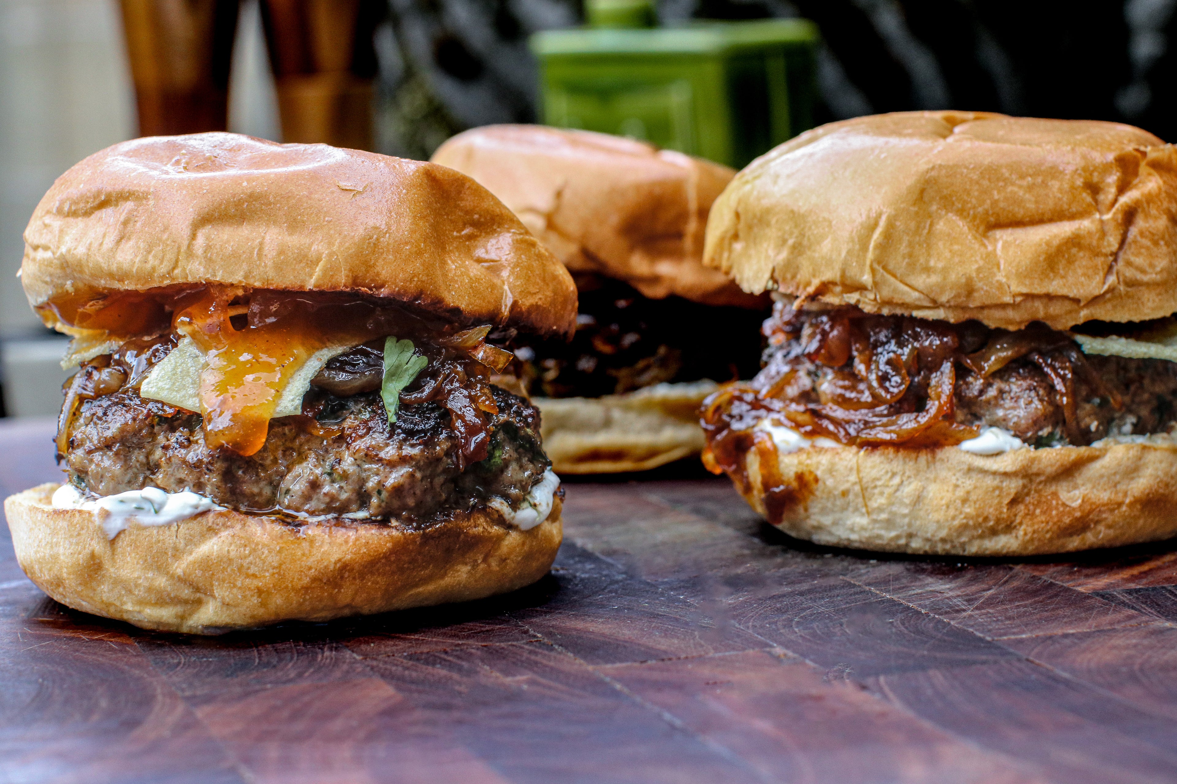 Three lamb burgers on buns with various toppings on a wooden surface.