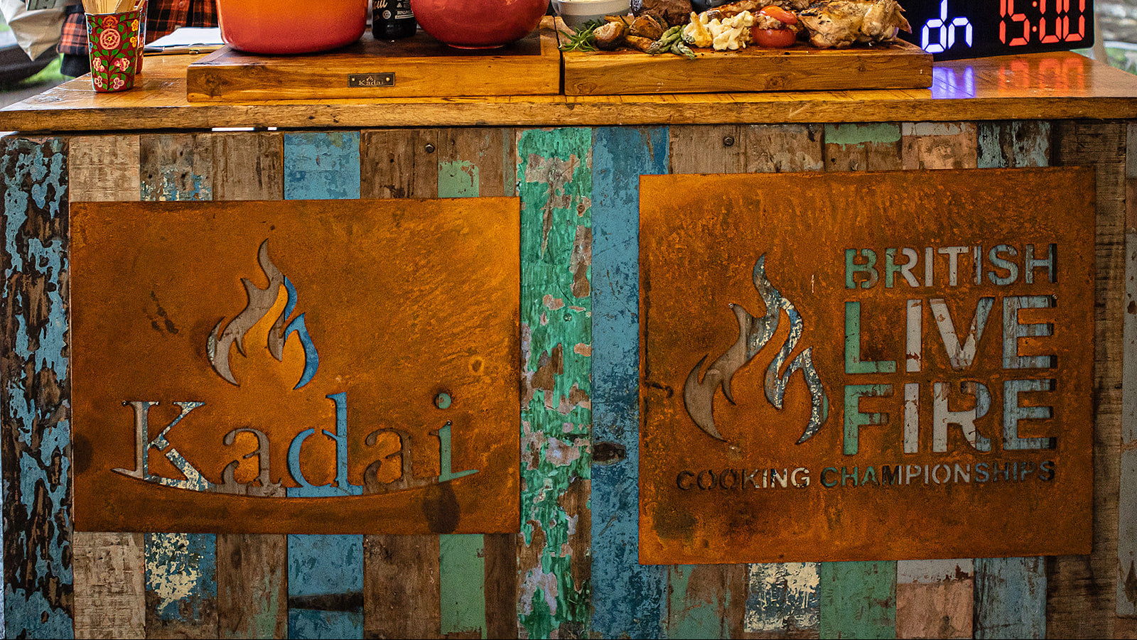 Wooden counter with 'Kadai' and 'British Live Fire Cooking Championships' signs, food items, and a digital clock.