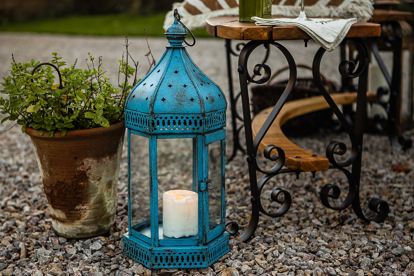 Blue lantern with a candle on a gravel surface, with a potted plant and outdoor furniture in the background.