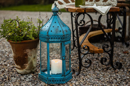 Blue lantern with a candle on a gravel surface, with a potted plant and outdoor furniture in the background.