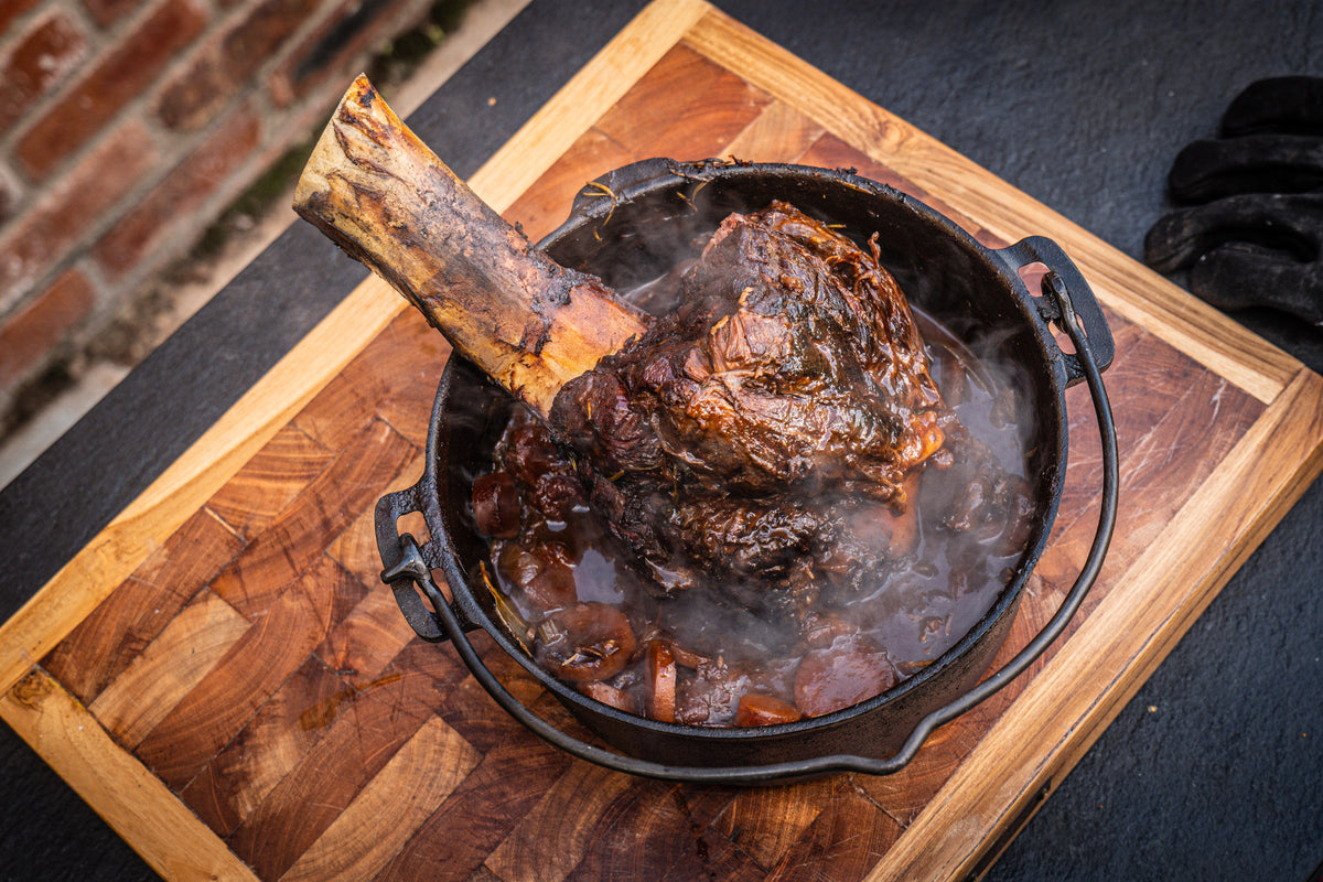 A large bone-in beef shank simmering in a dark, rich sauce inside a black cast iron dutch oven, presented on a wooden cutting board