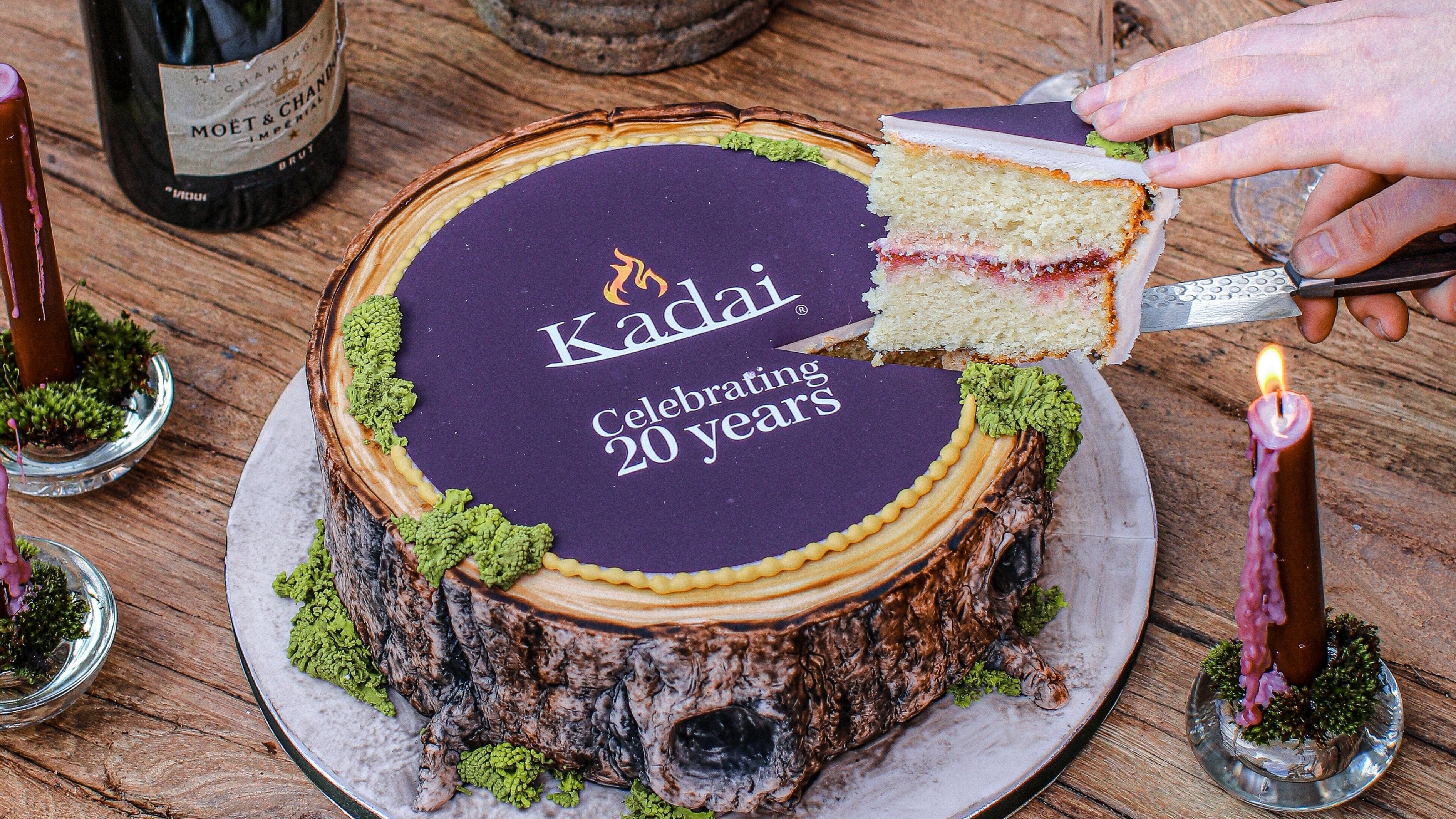 Kadai cake on a table with a bottle of champagne and someone taking a slice.