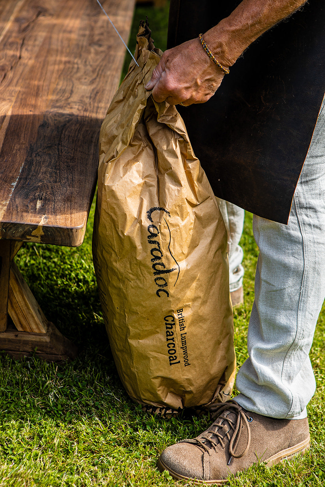 Person holding a brown paper bag with 'Caradoc' charcoal branding on grass next to a wooden table.
