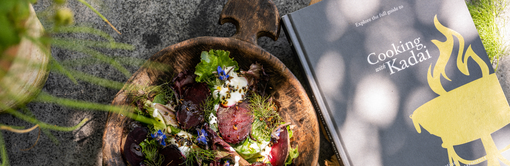 Wooden bowl with salad and a book titled 'Cooking with Kadau' on a textured surface with plants.