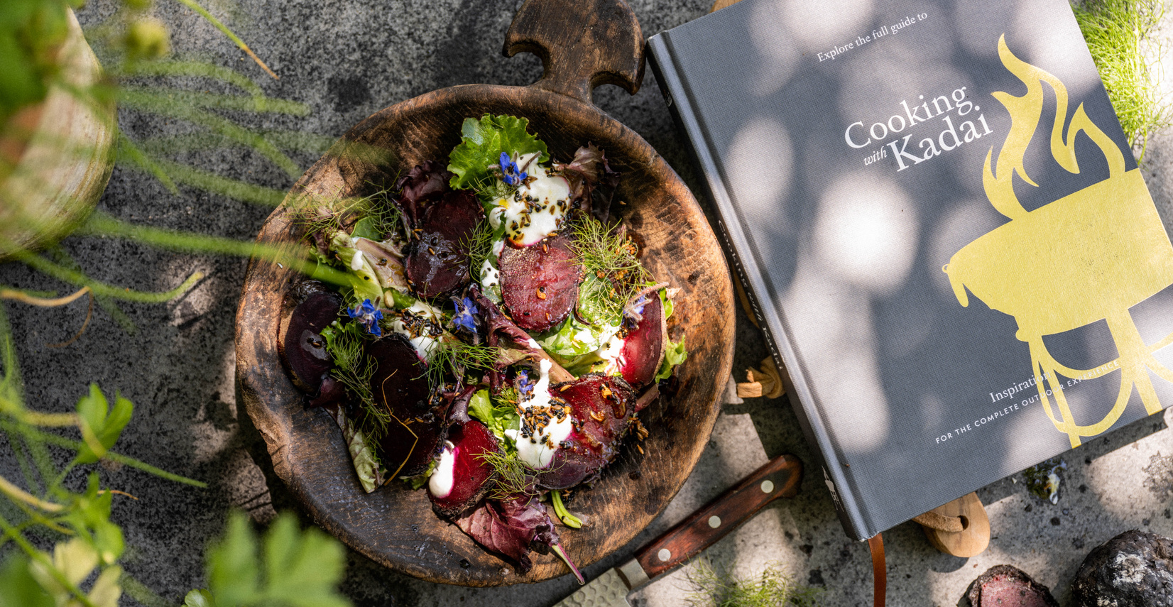 Wooden bowl with salad and a book titled 'Cooking with Kadai' on a textured surface.