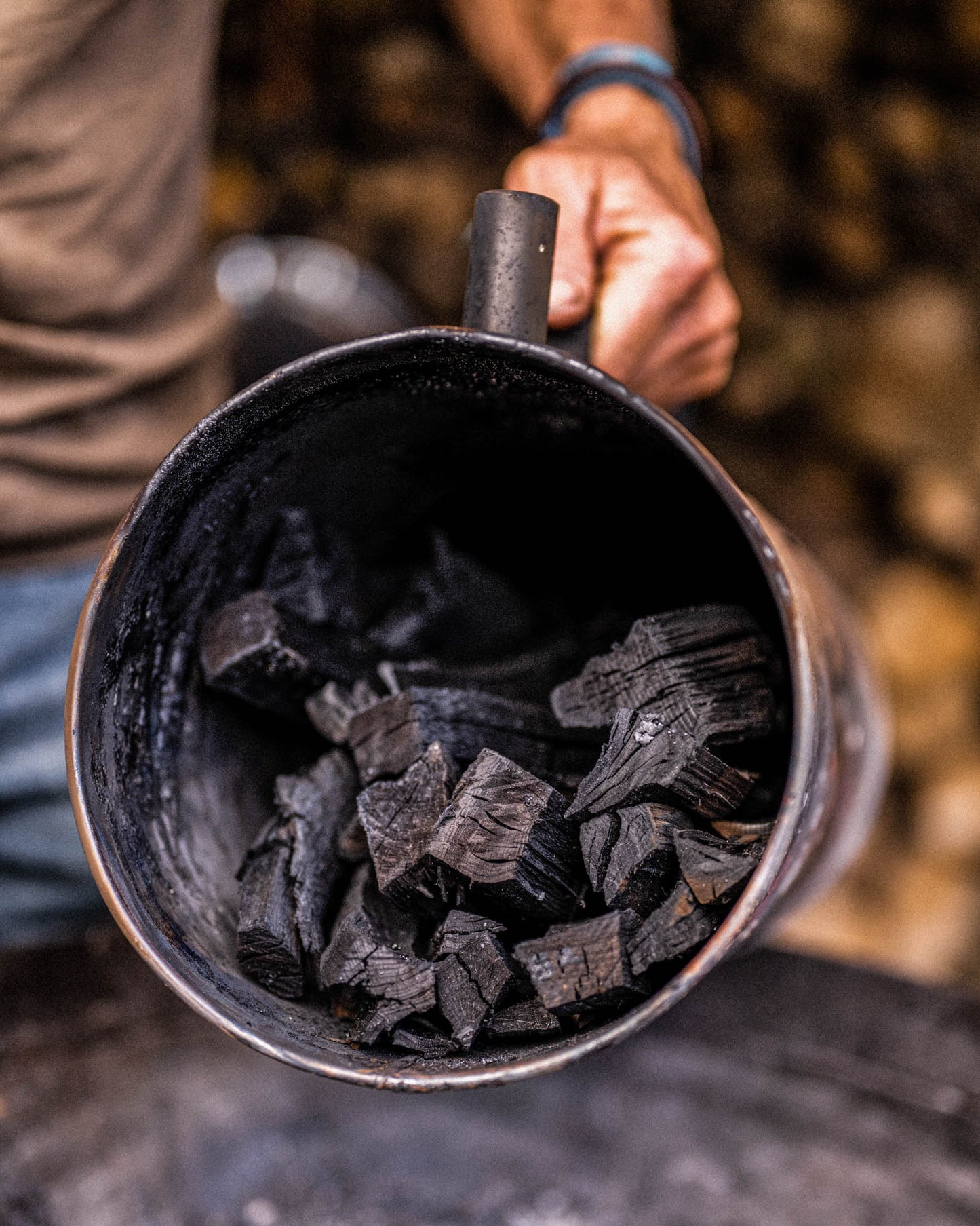 Close-up of a charcoal maker container filled with freshly made black charcoal pieces, held by hand against a blurred background of stacked firewood.