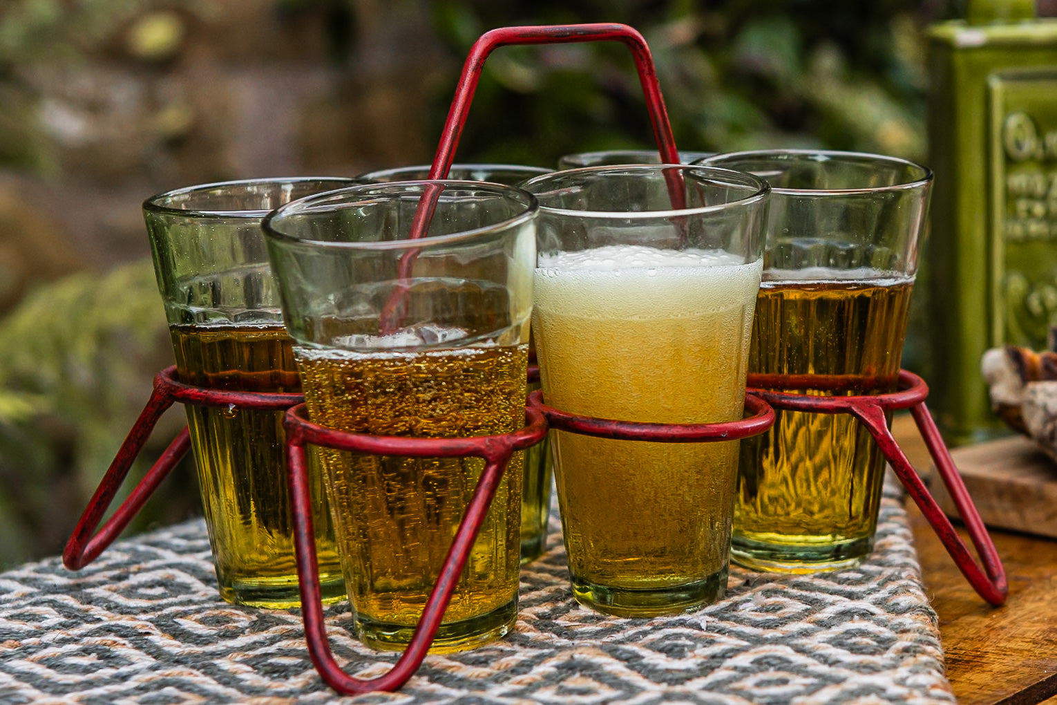 Glasses of beer in a red metal holder on a patterned table, outdoors.