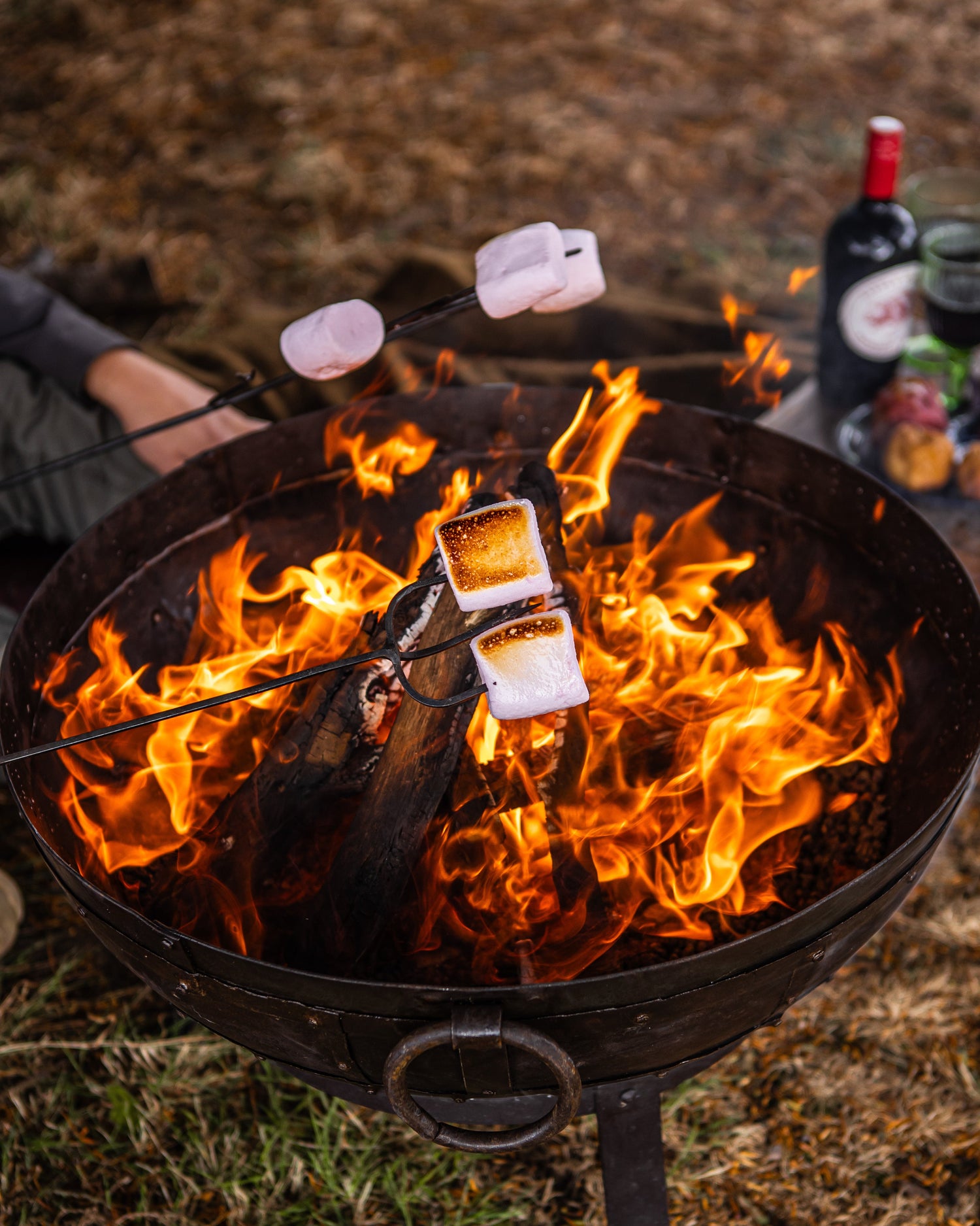 Person roasting marshmallows over a campfire with bottles and food in the background