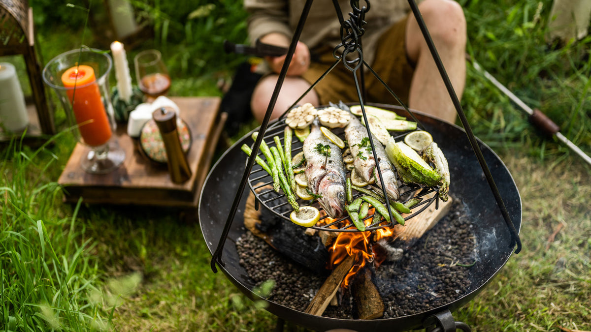 Outdoor cooking scene with fish and vegetables on a grill over a fire, surrounded by nature.