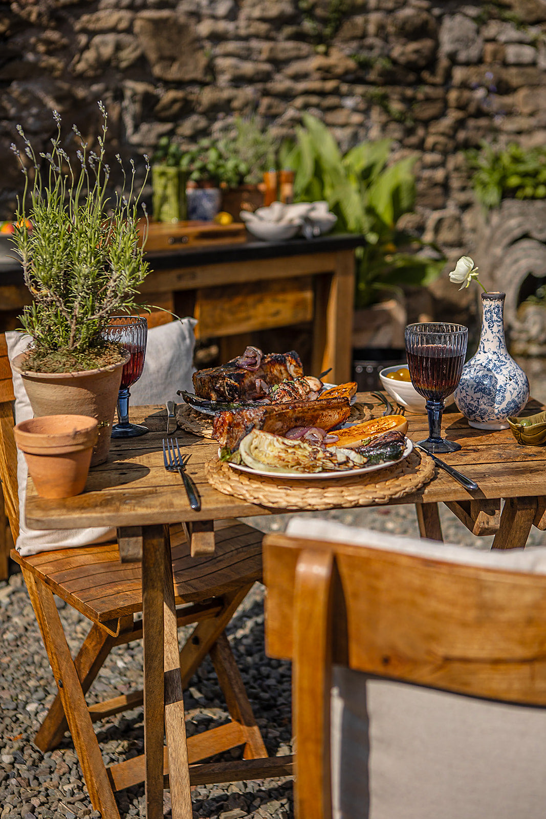 Rustic outdoor dining setup with wooden café chairs surrounding a table filled with grilled food, wine glasses, and garden décor