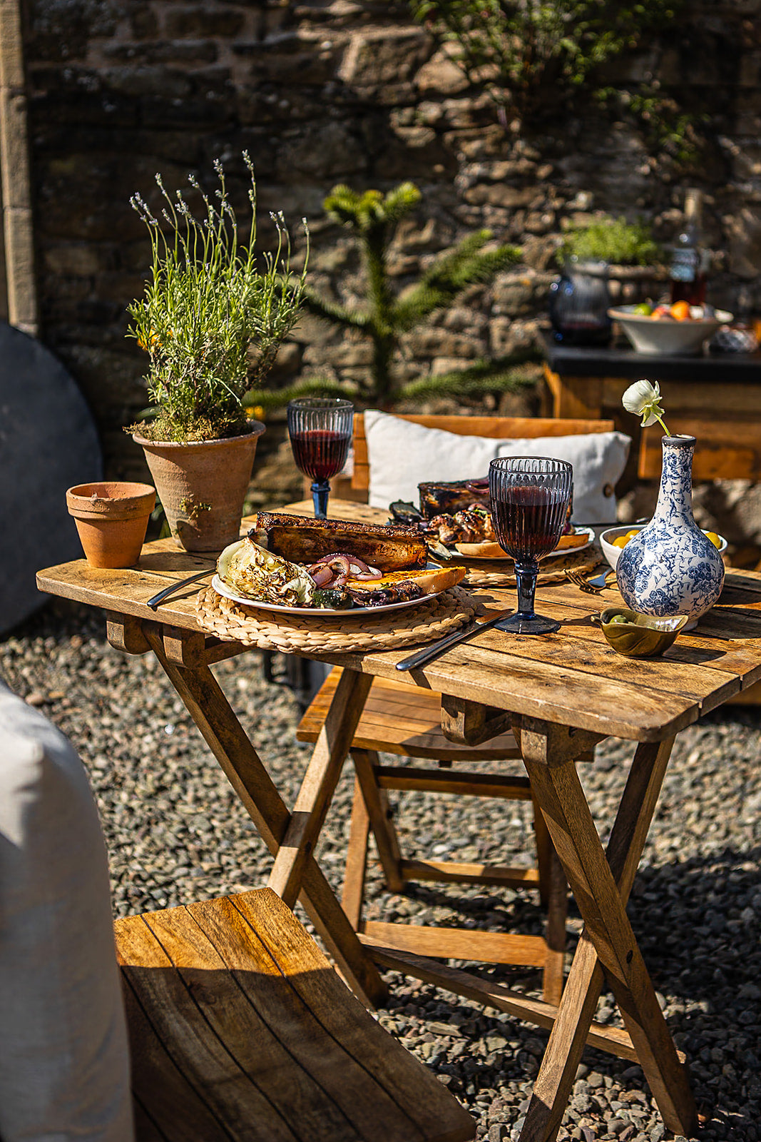 Rustic wooden café table styled for outdoor dining with grilled food, wine glasses, and decorative tableware in a sunlit courtyard