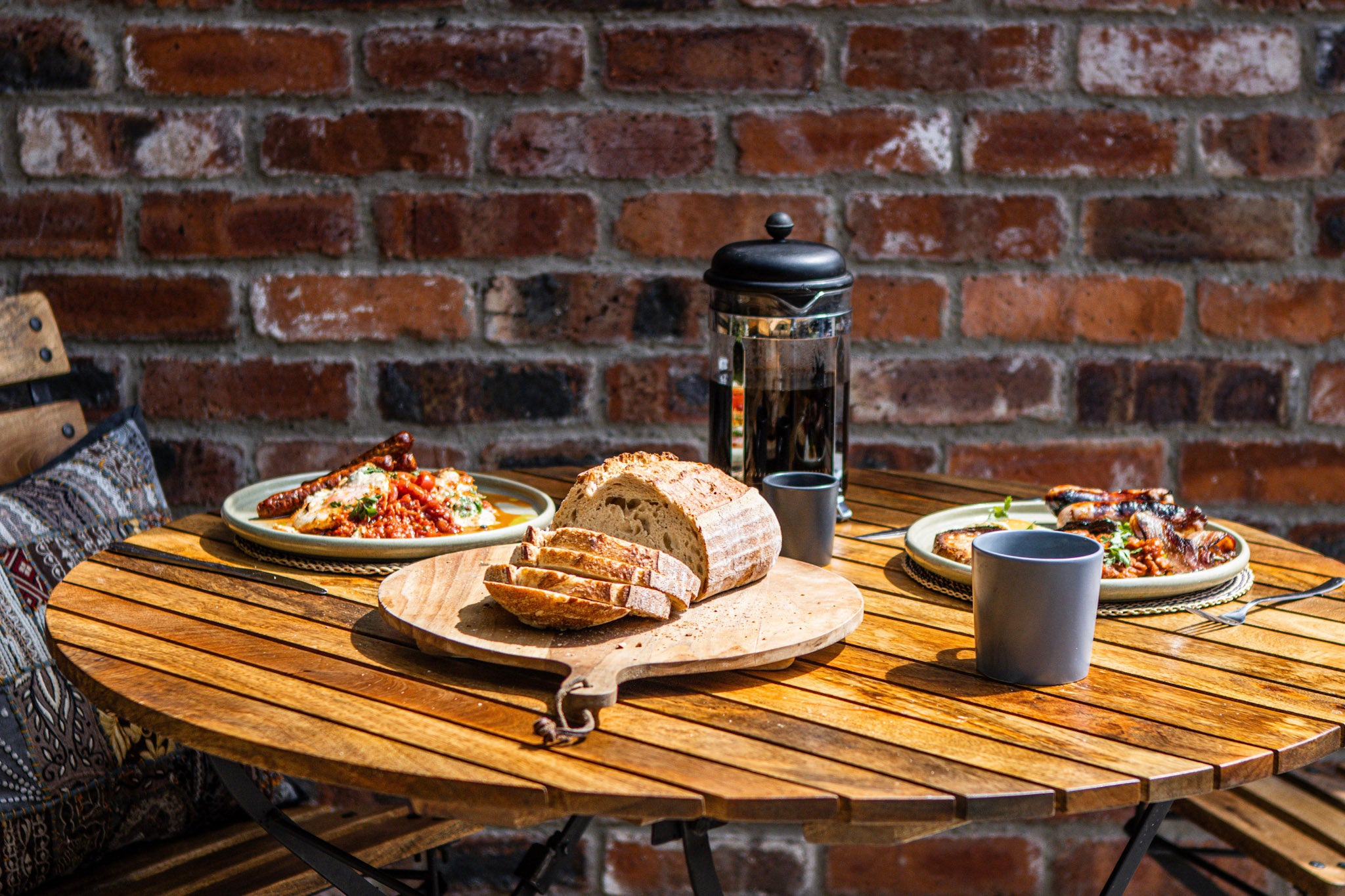 A rustic wooden round table set against a red brick wall, featuring a loaf of bread on a wooden board, a black French press coffee maker, plates of food, and gray mugs.
