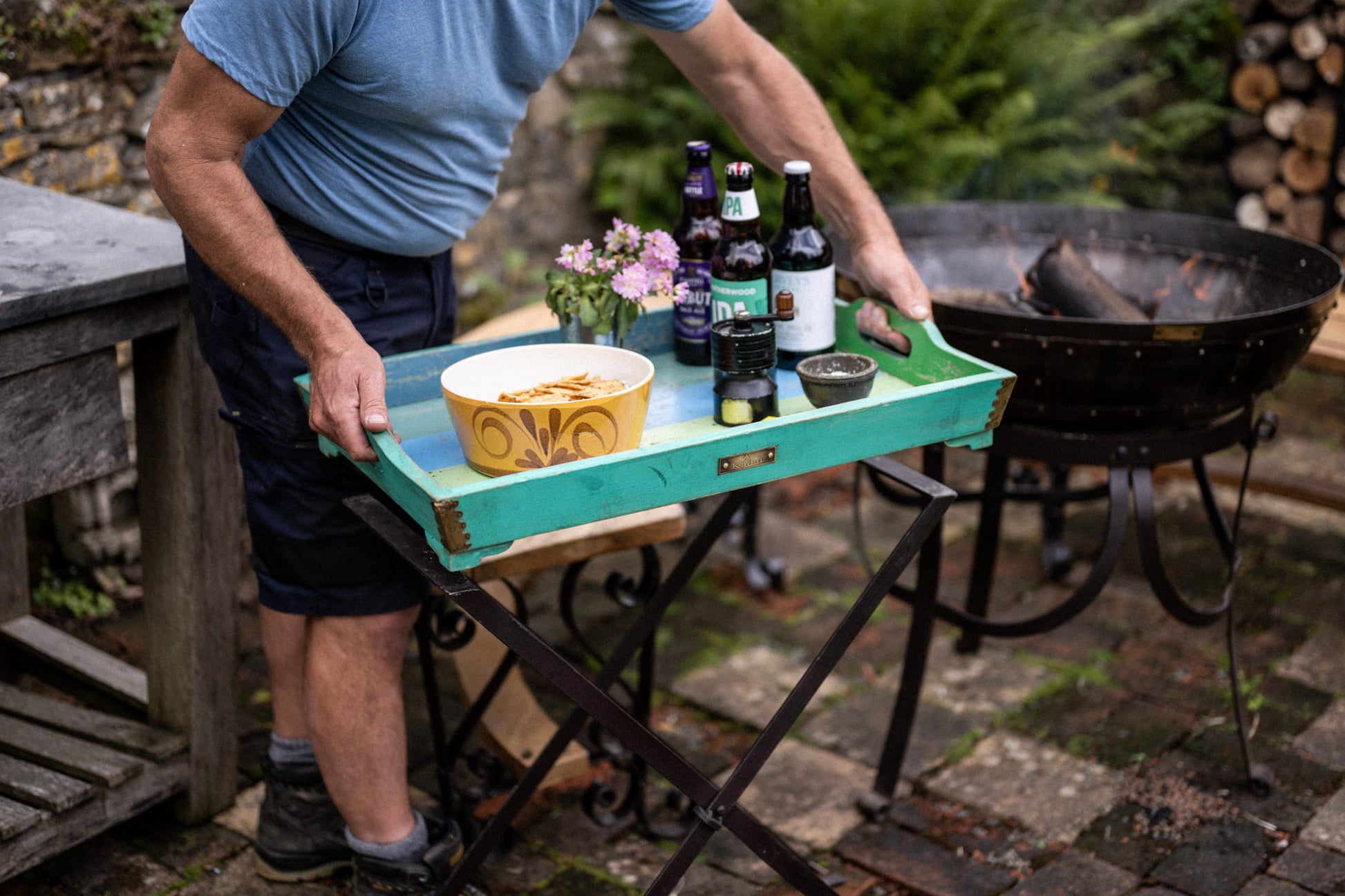 Person setting down a turquoise folding tray table with drinks, snacks and flowers beside a rustic outdoor fire bowl on a stone patio.