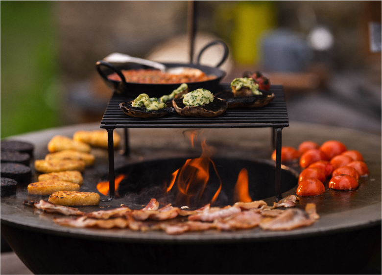 Kadai firepit grilling bacon, tomatoes, and mushrooms with elevated cooking rack outdoors