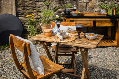 Rustic wooden café chairs arranged around an outdoor dining table with wine glasses, ceramics, and garden plants in a stone courtyard