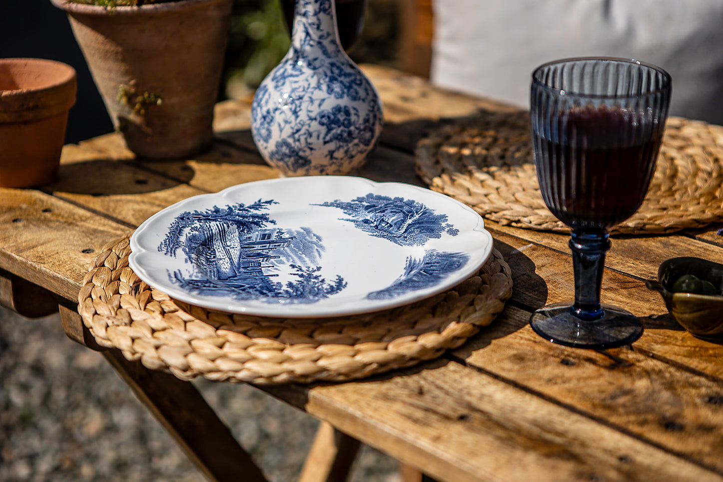 Rustic wooden café table set with blue-and-white ceramic tableware, woven placemats, and a dark goblet for outdoor dining