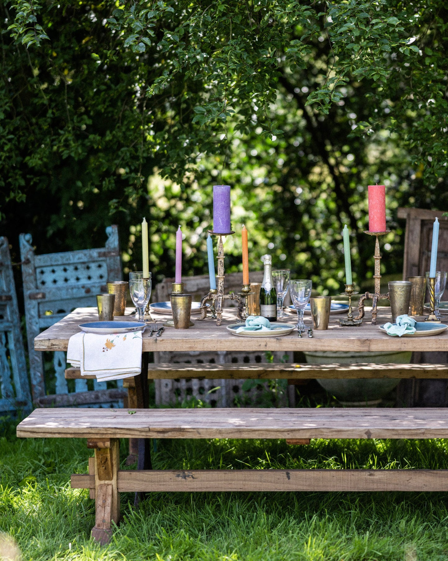Rustic teak garden bench positioned beside an outdoor dining table styled with colourful candles, brass tableware and glassware in a lush garden.