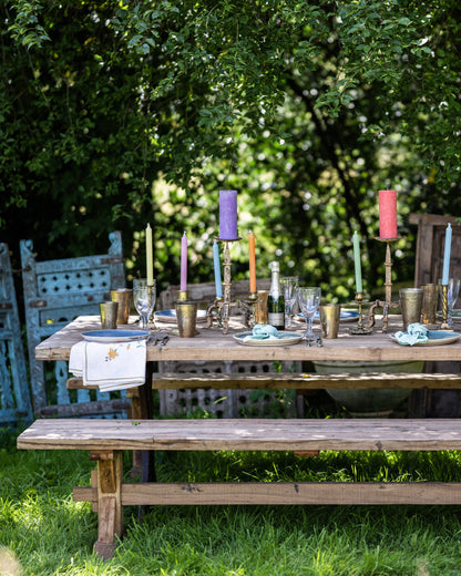 Rustic teak garden bench positioned beside an outdoor dining table styled with colourful candles, brass tableware and glassware in a lush garden.