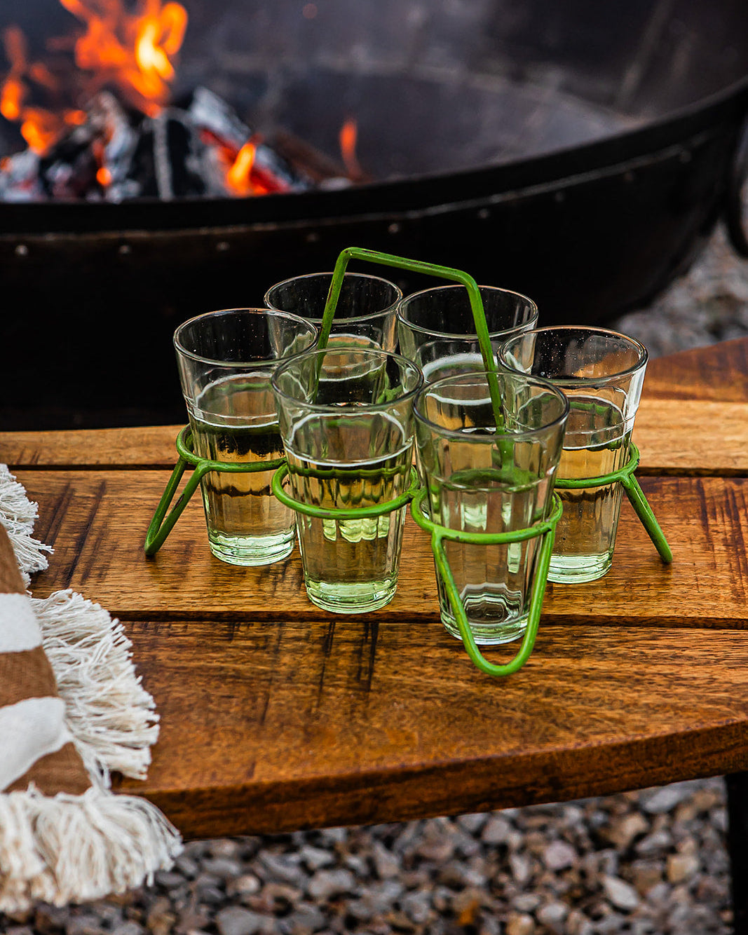 Set of chai glasses in green metal holder on wooden bench beside outdoor fire pit