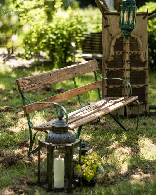 Wooden garden bench with metal frame on grass, surrounded by lanterns and greenery.