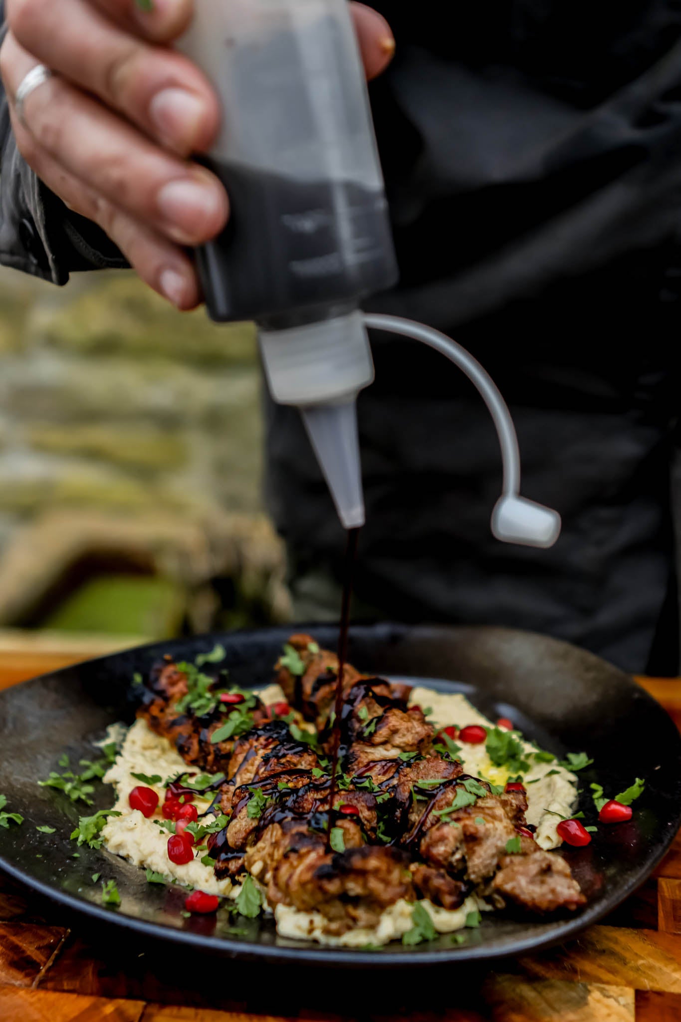 Person drizzling a dressing on a plate of hummus and grilled lamb skewers