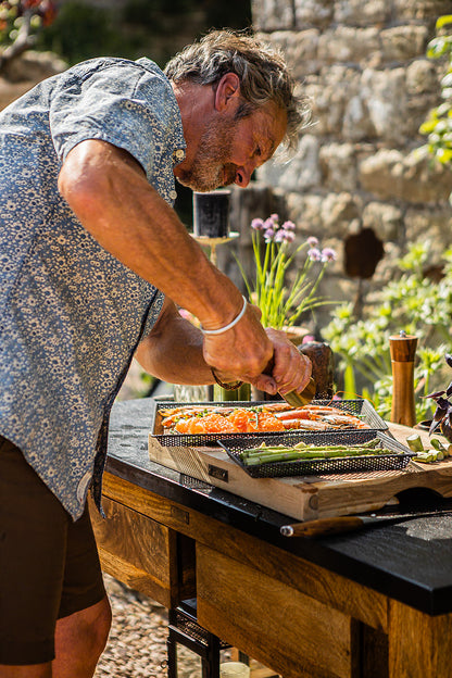 Man preparing food on a grill outdoors with a stone wall and plants in the background