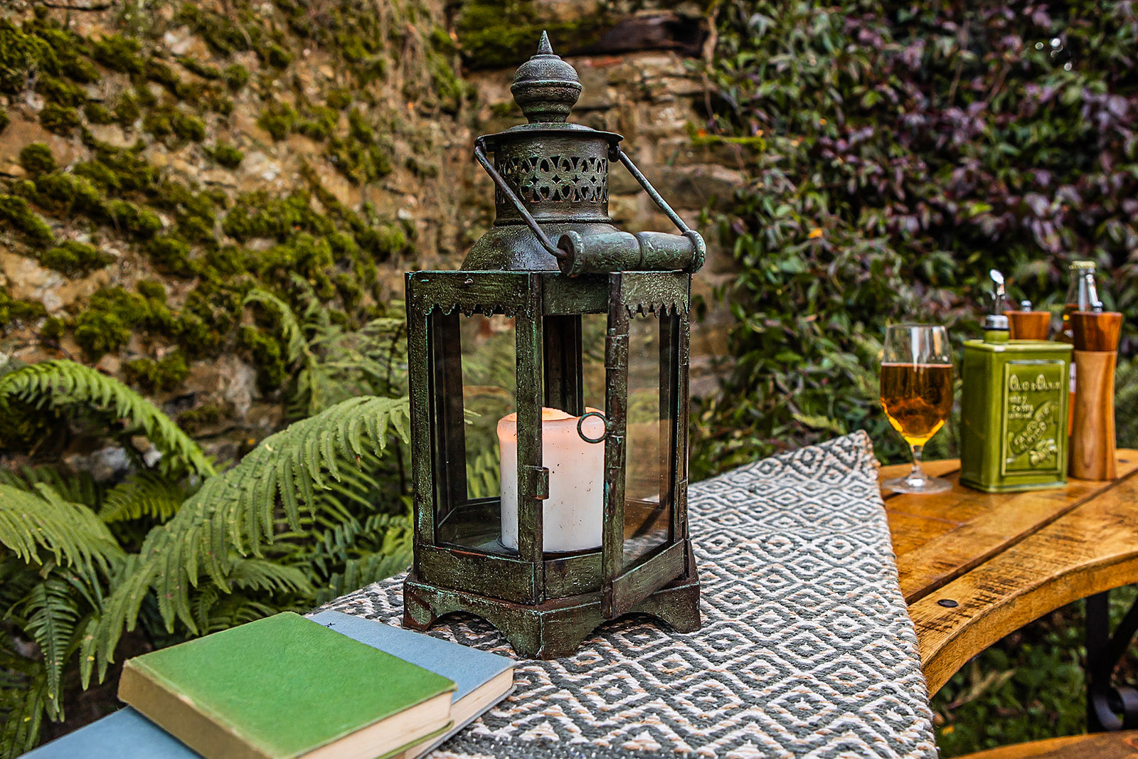Decorative lantern with a candle on a table outdoors, surrounded by plants and a glass of wine.