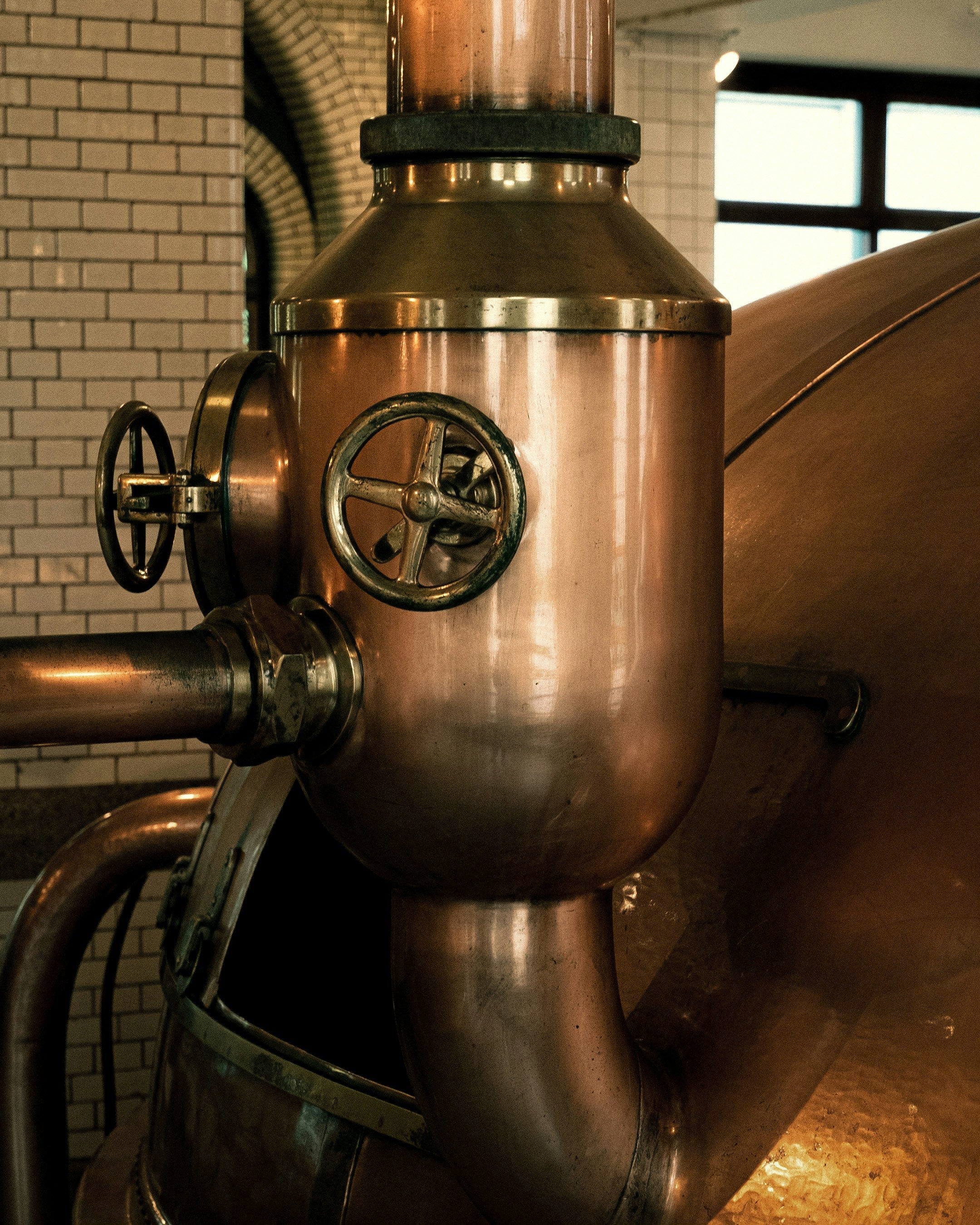 Copper still in a distillery setting with brick wall and windows.
