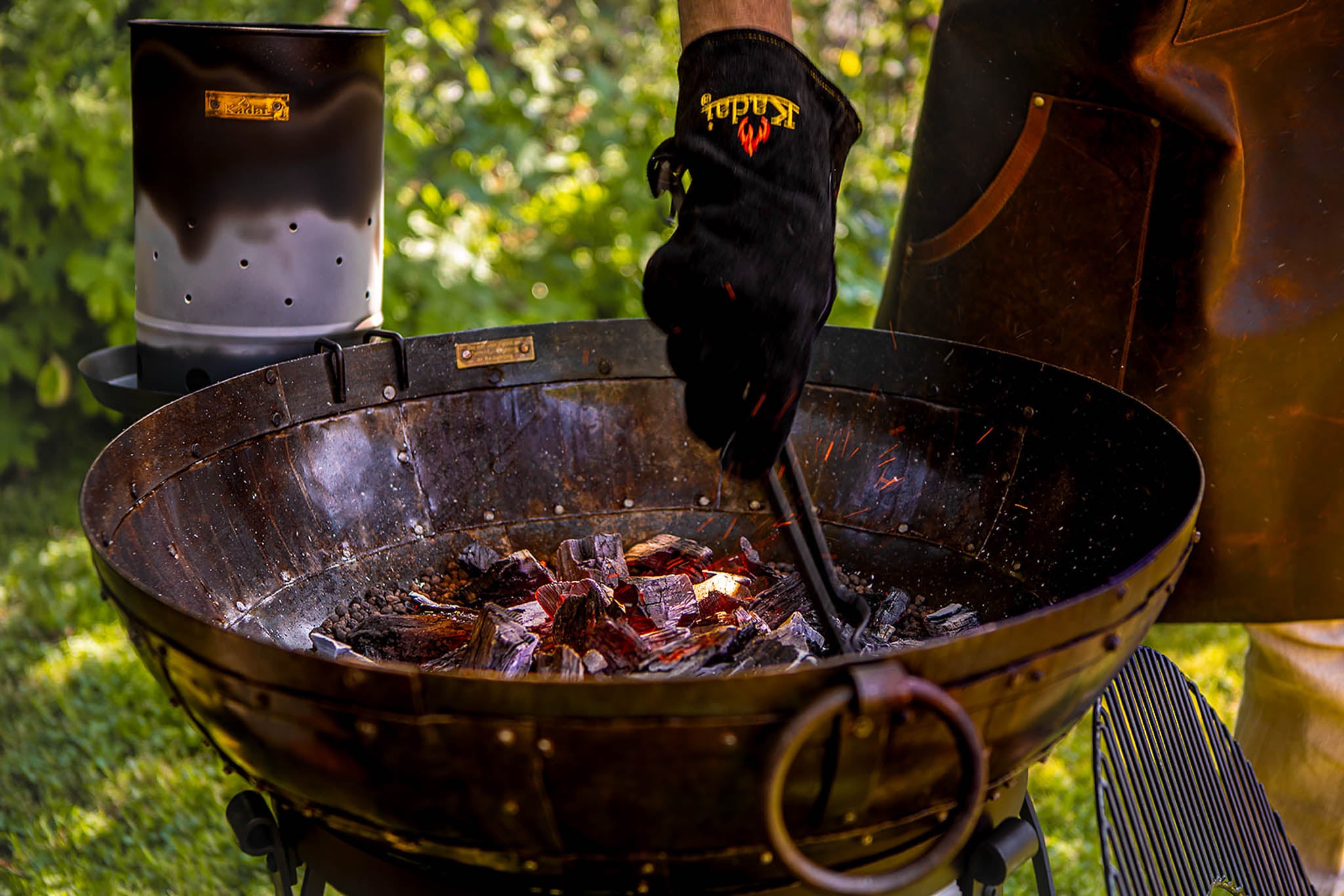 Person grilling with a barbecue and tongs, wearing branded gloves.