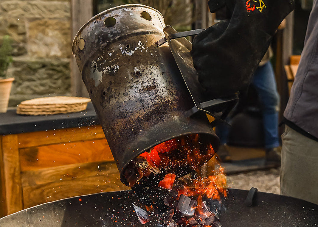 Person pouring hot coals from a metal container into a barbecue grill.
