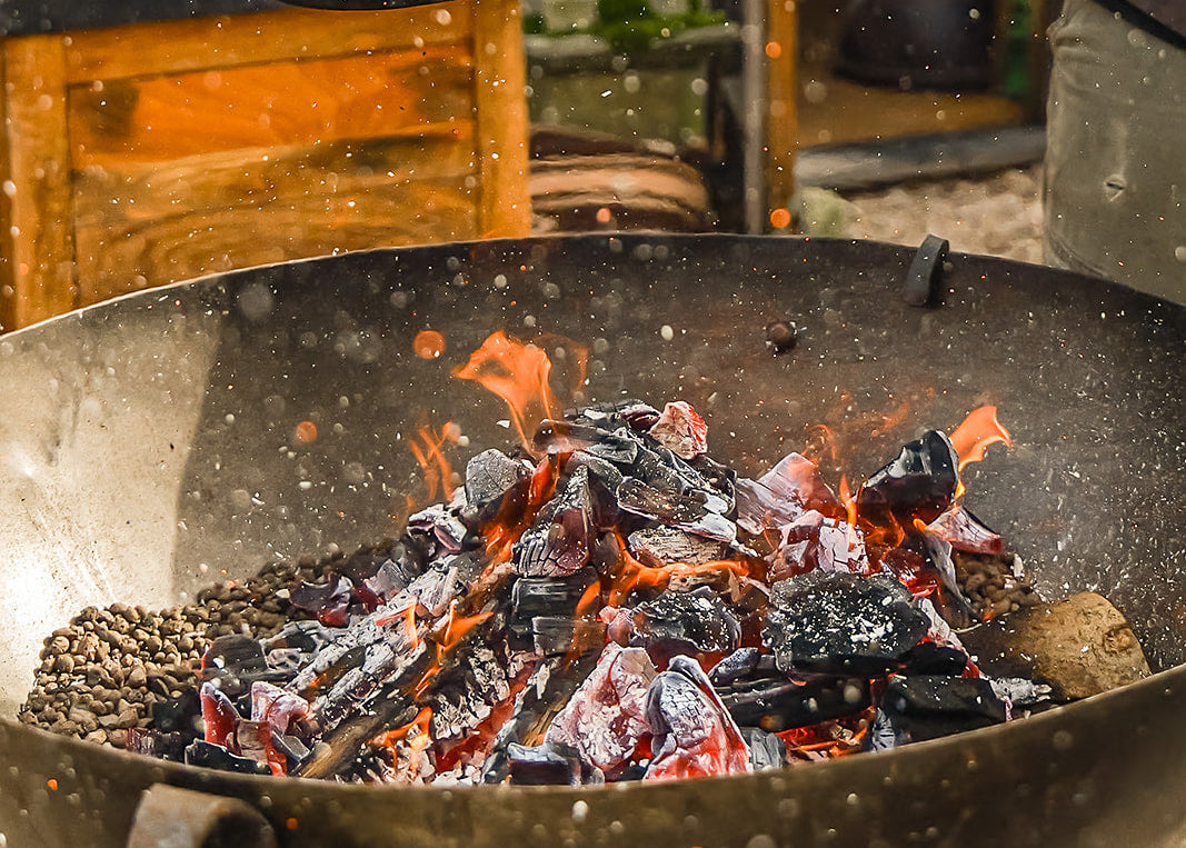 Person using a metal container to add charcoal to a barbecue grill outdoors.