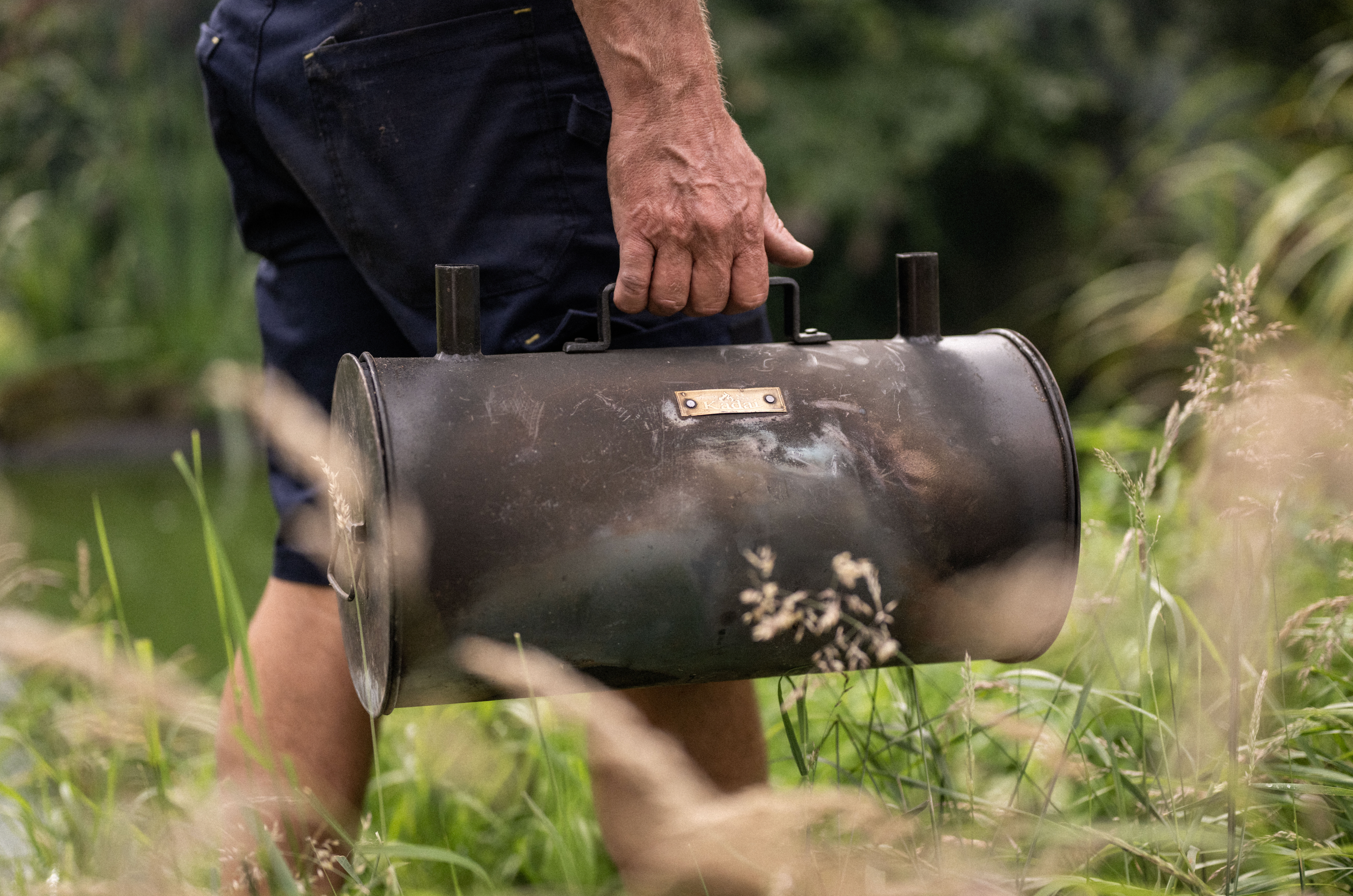 Person carrying a large cylindrical metal charcoal maker in long grass.