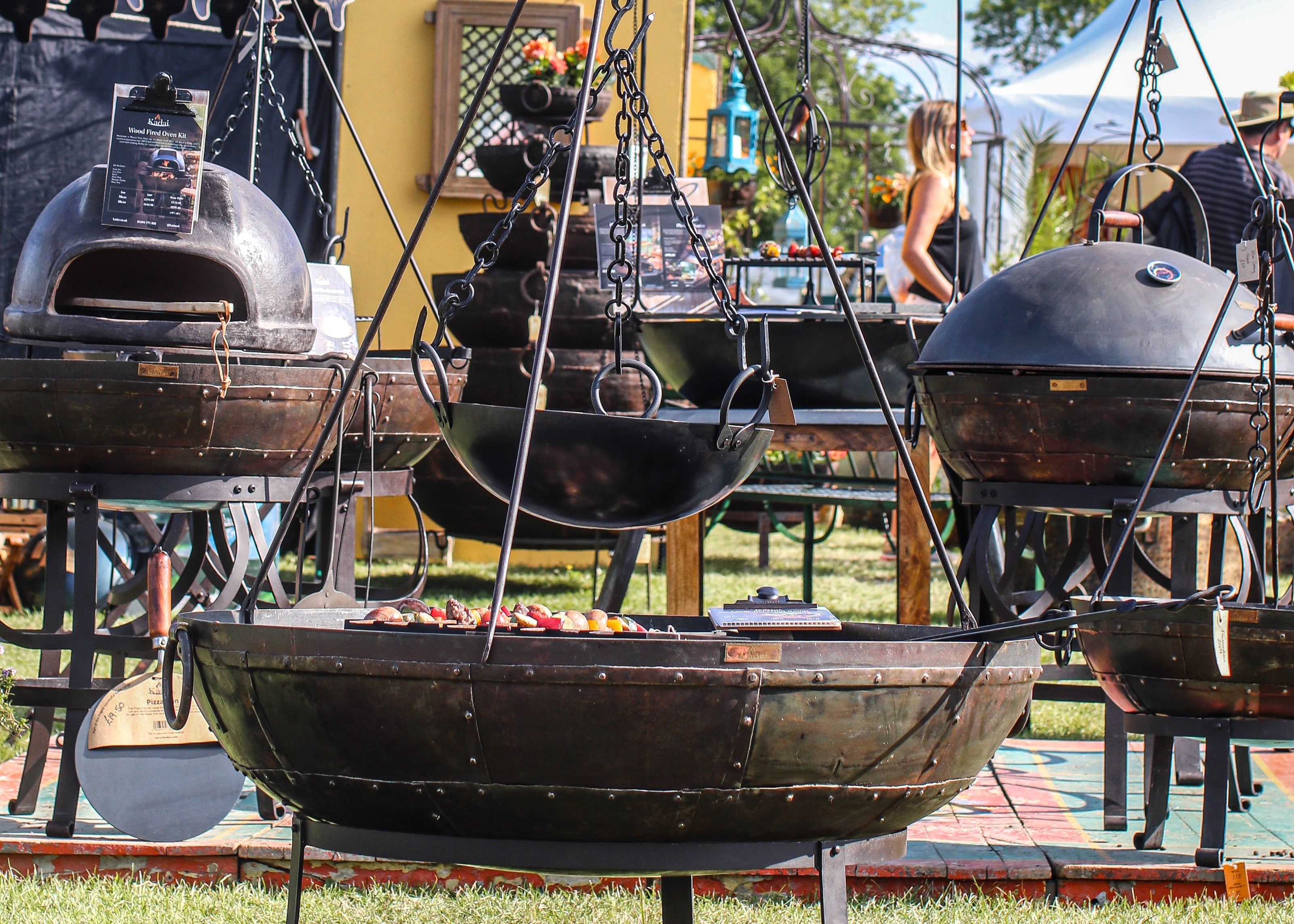 Outdoor setting with large metal kadais and grills on a grassy area.