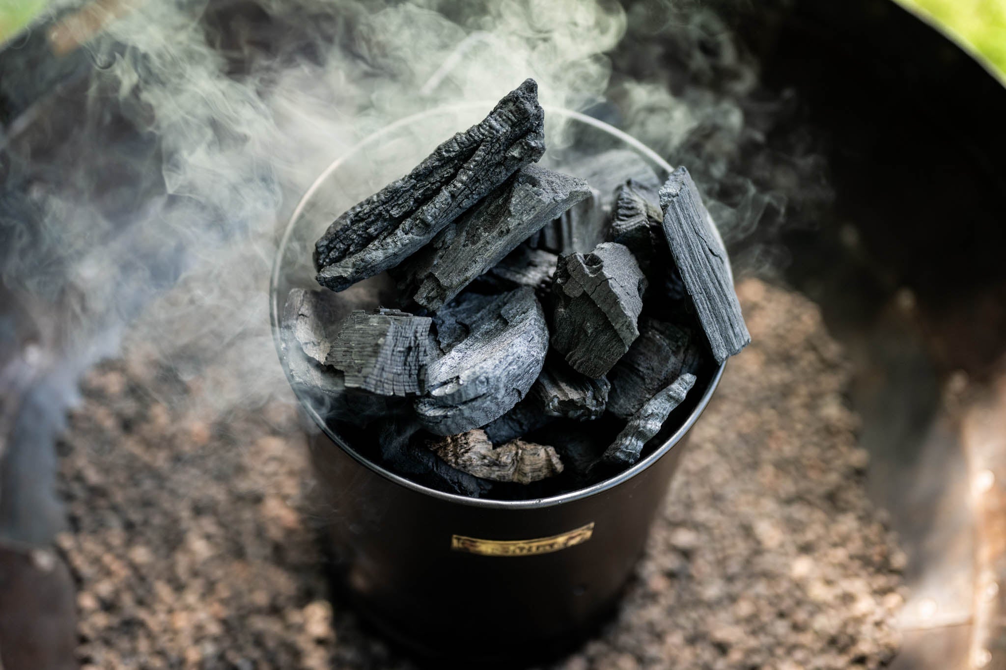 Charcoal briquettes in a Kadai Charcoal Starter with smoke rising, on a blurred background.