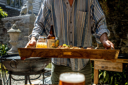 A rustic wooden serving tray held by a person outdoors, styled with a bottle of beer, a glass, and a small food platter. The scene is set in a garden or patio environment with a fire pit and stone walls in the background.