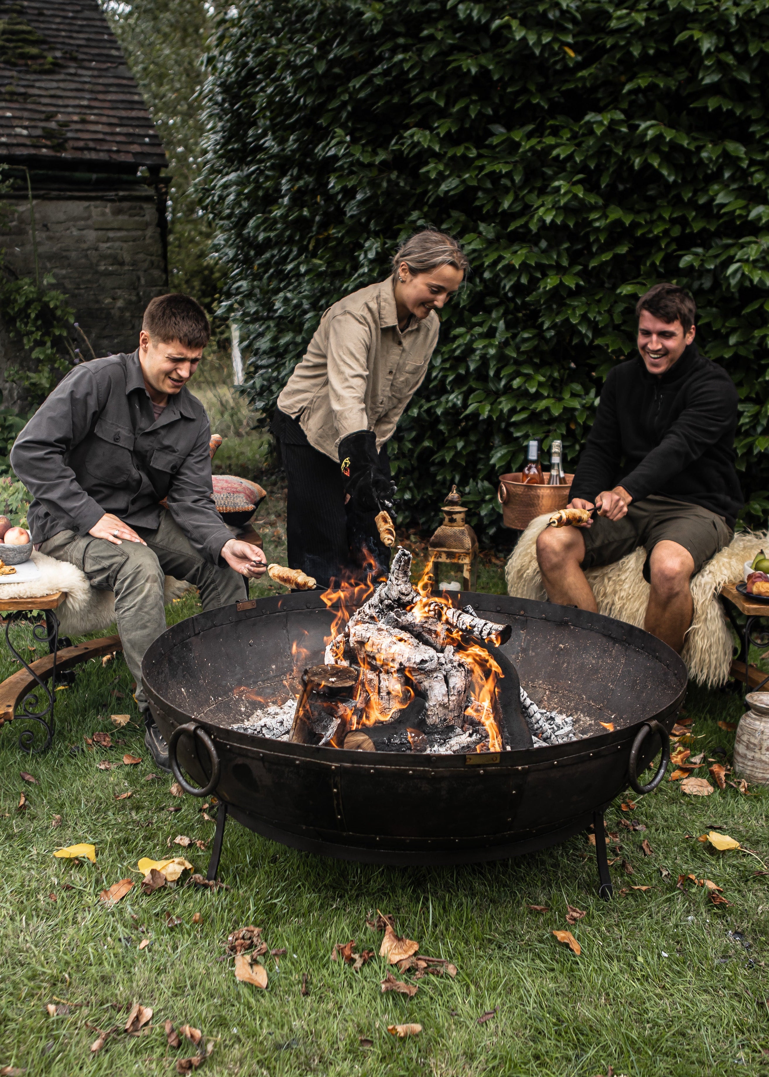 Three people around a fire pit in an outdoor setting with greenery.