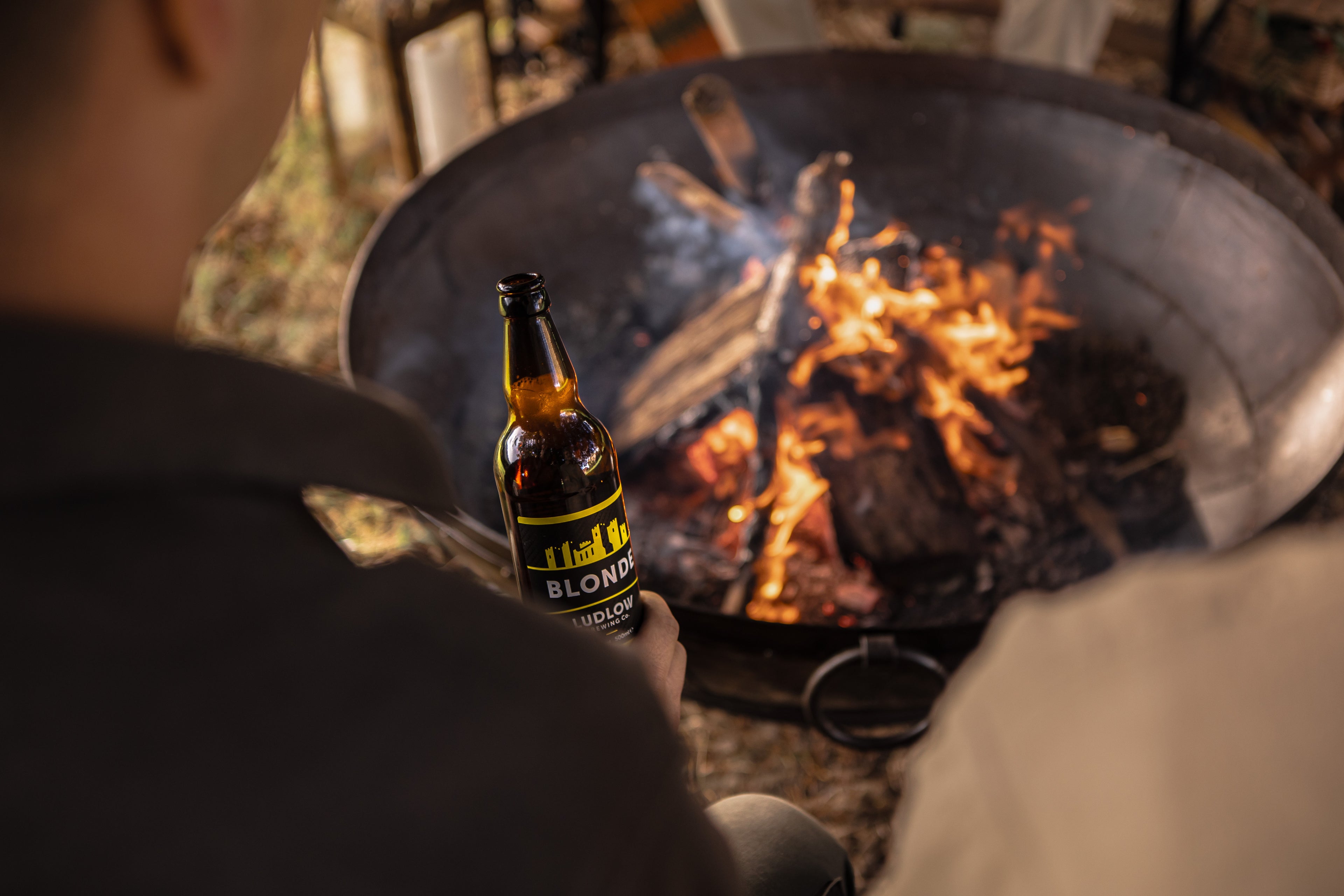 Person holding a bottle of beer by a kadai fire pit with flames