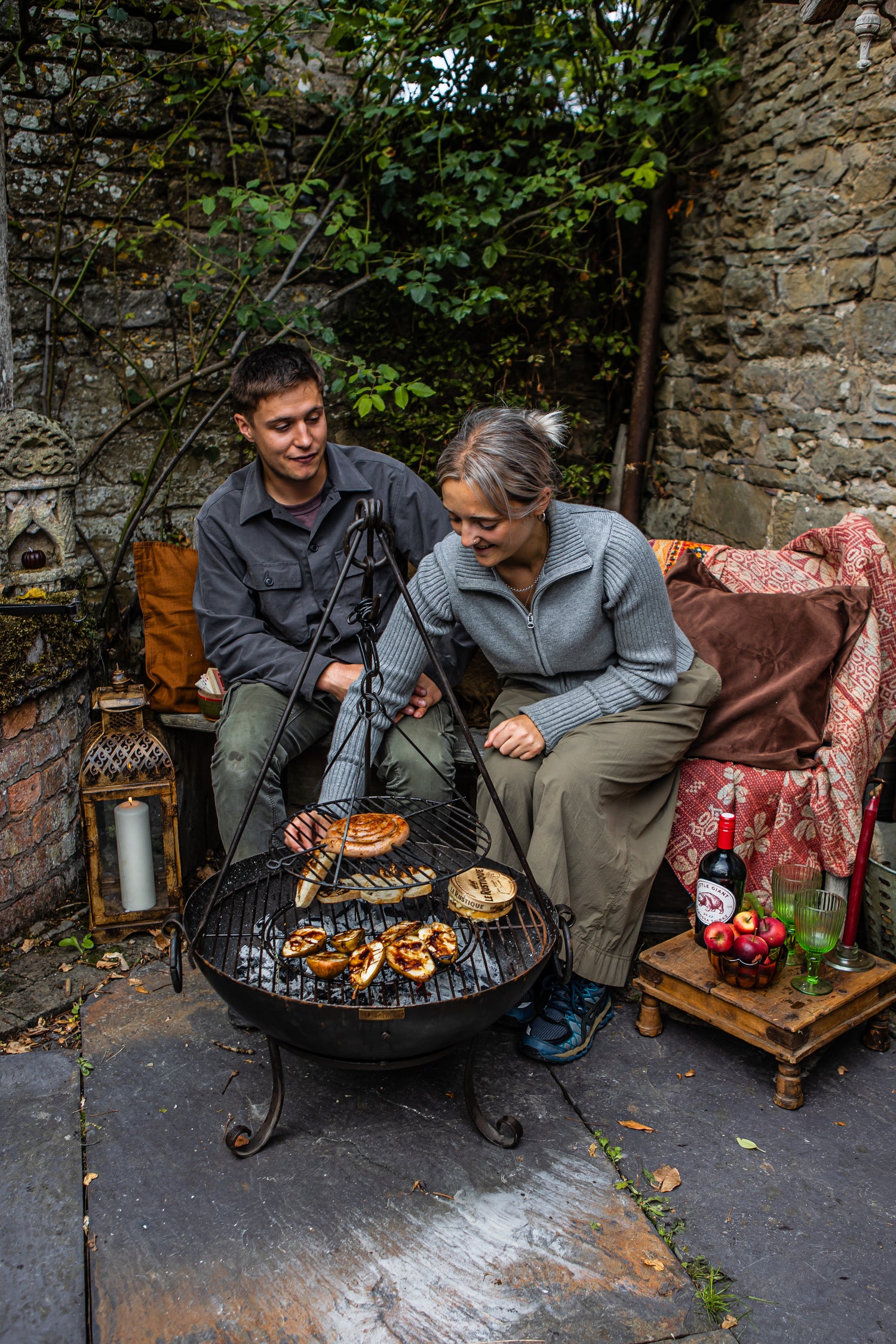 Two people grilling food on kadai firebowl outdoors near a stone well with a rustic setting.